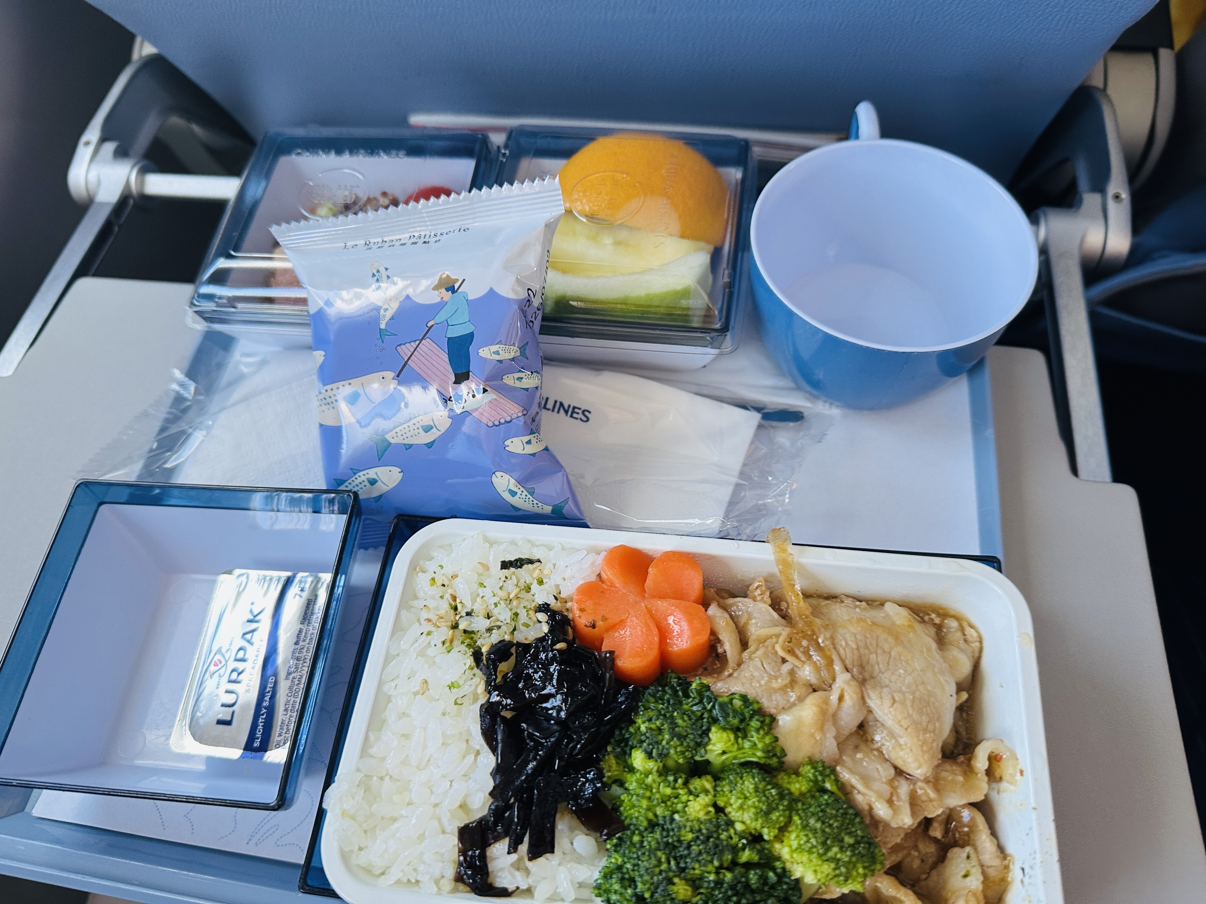 飛機餐的餐盤，主餐是照燒豬肉配白飯、綠花椰菜與紅蘿蔔，旁邊有水果盒、麵包、奶油與一杯水