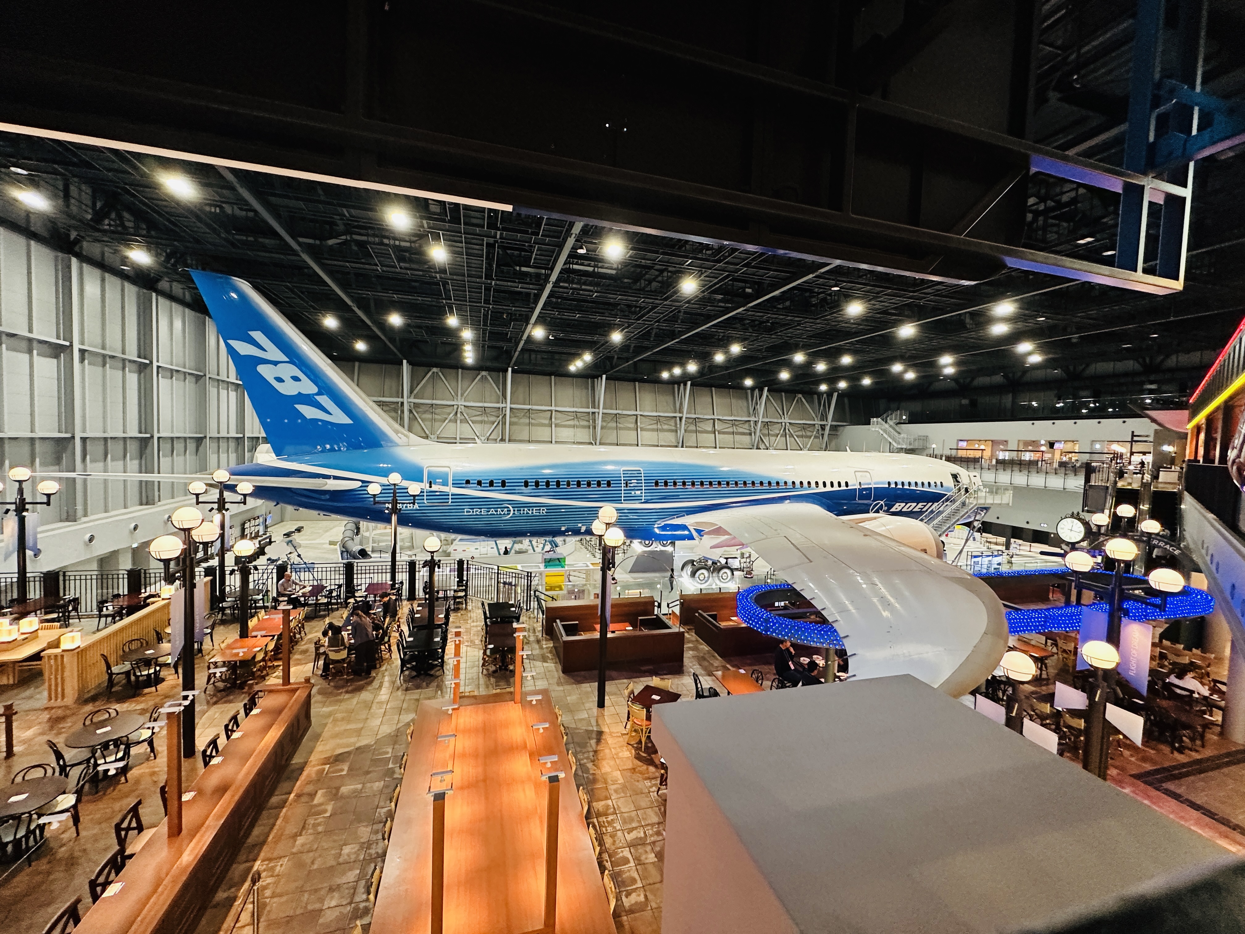 航空博物館內展示的波音 787 夢幻客機，藍白塗裝的機身停在挑高的機棚裡，下方是餐飲區的桌椅