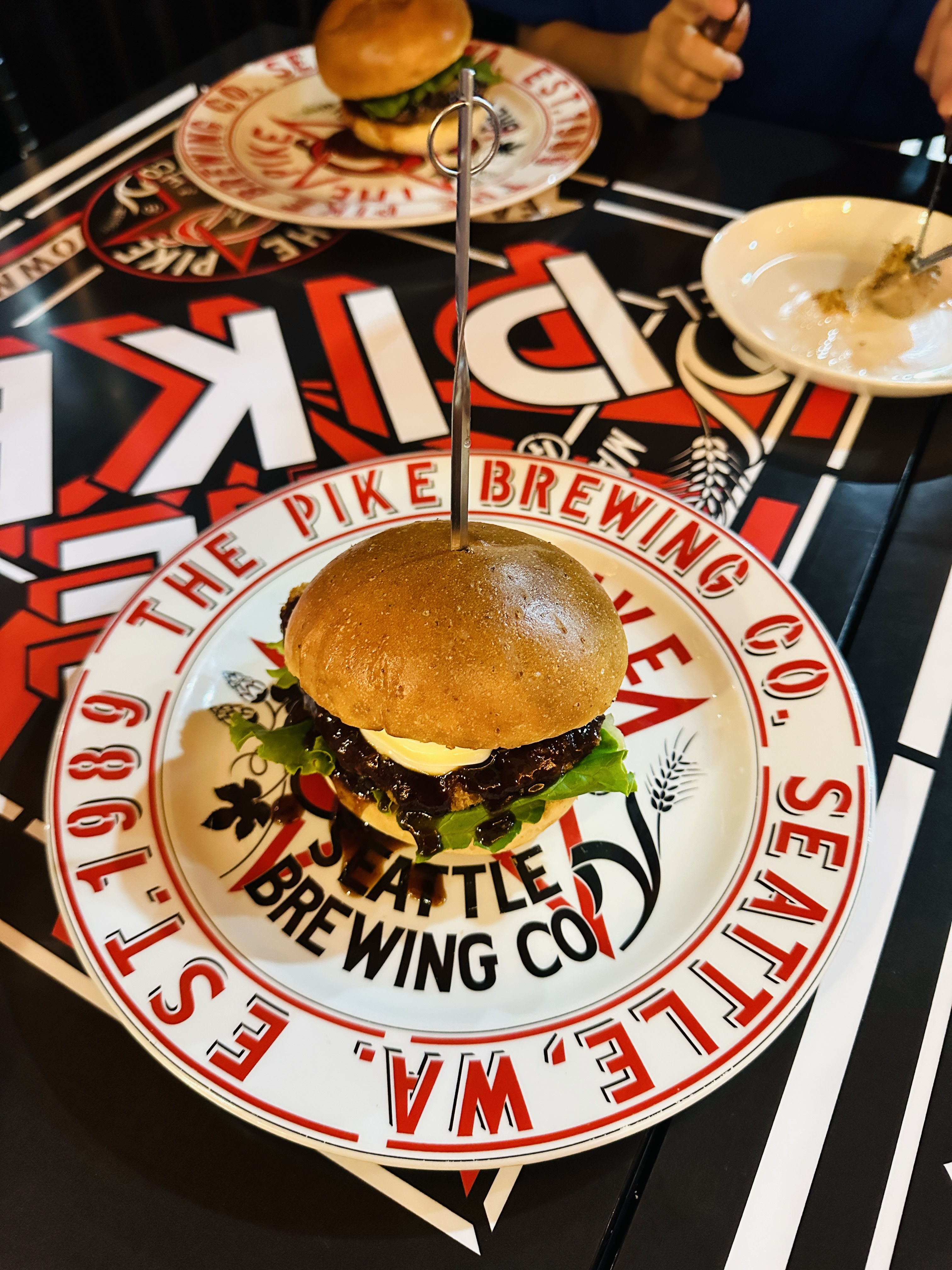白色圓盤上的漢堡，插著金屬叉，盤緣印著 THE PIKE BREWING CO. SEATTLE 的紅色字樣