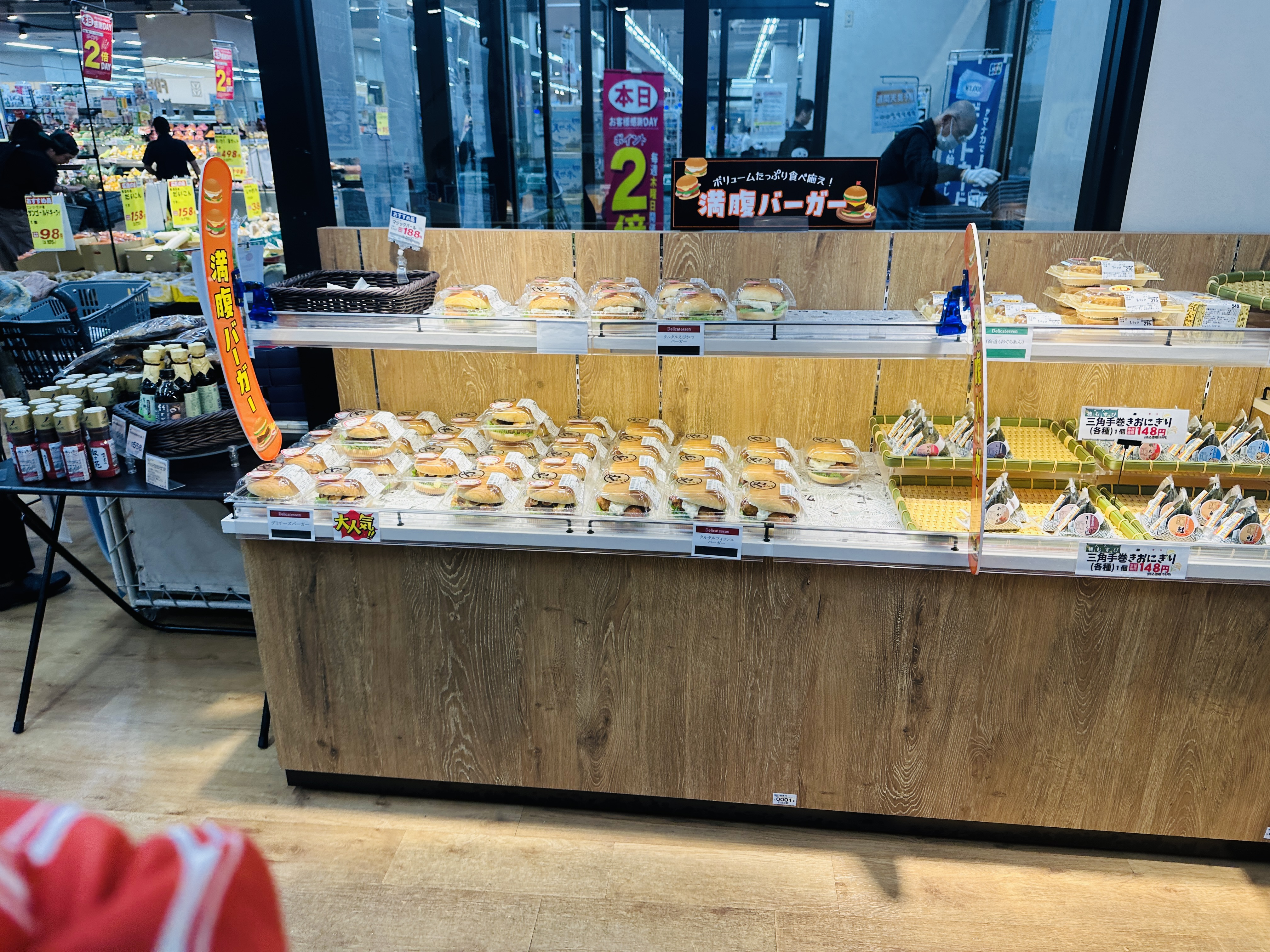 超市的麵包櫃，木質層架上整齊排著各式三明治與漢堡，後方貼著特價海報