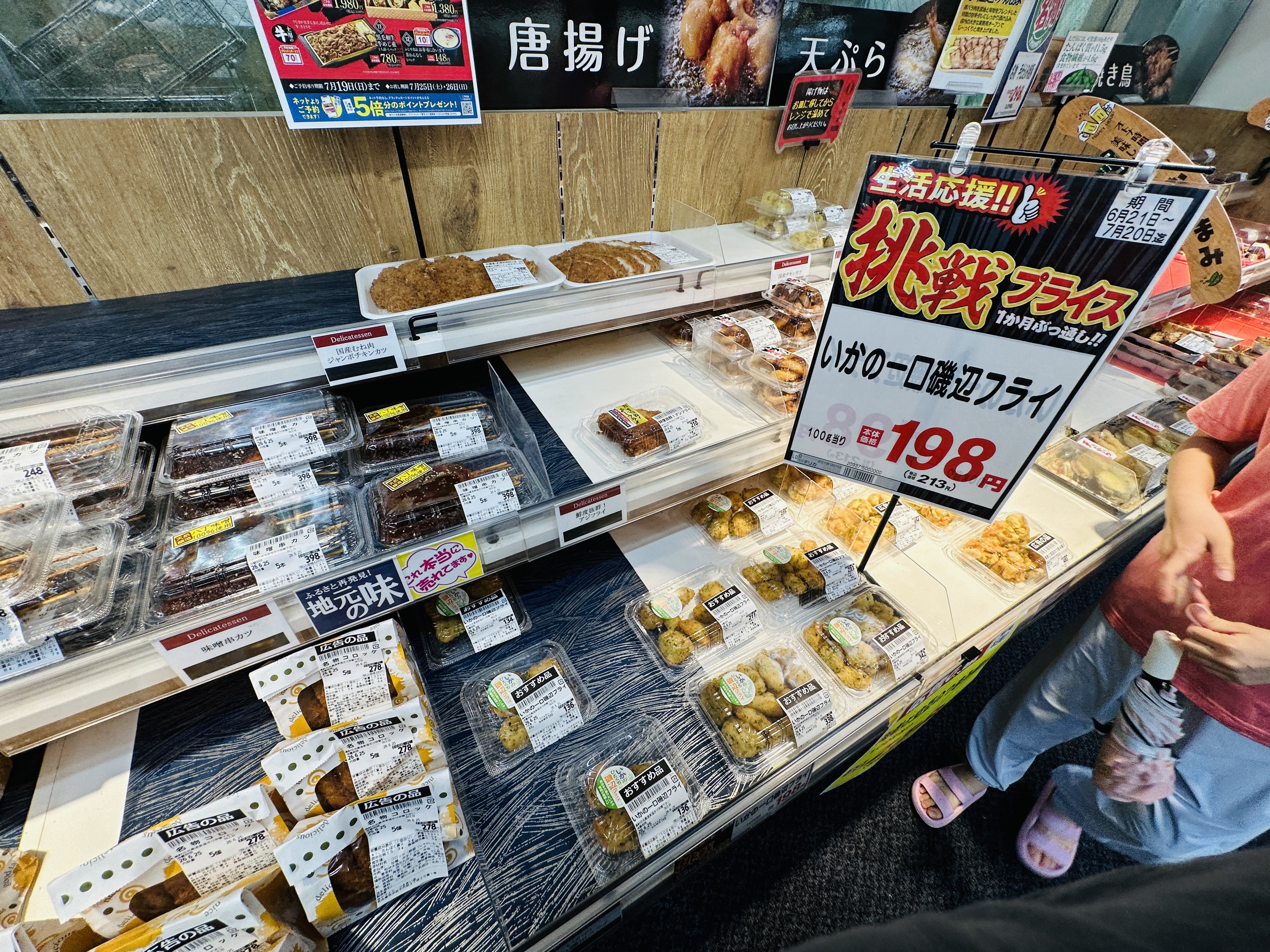 超市熟食區的冷藏櫃，陳列著炸物與便當，上方掛著「唐揚げ」「天ぷら」的看板，中央立著一張 198 圓的特價牌