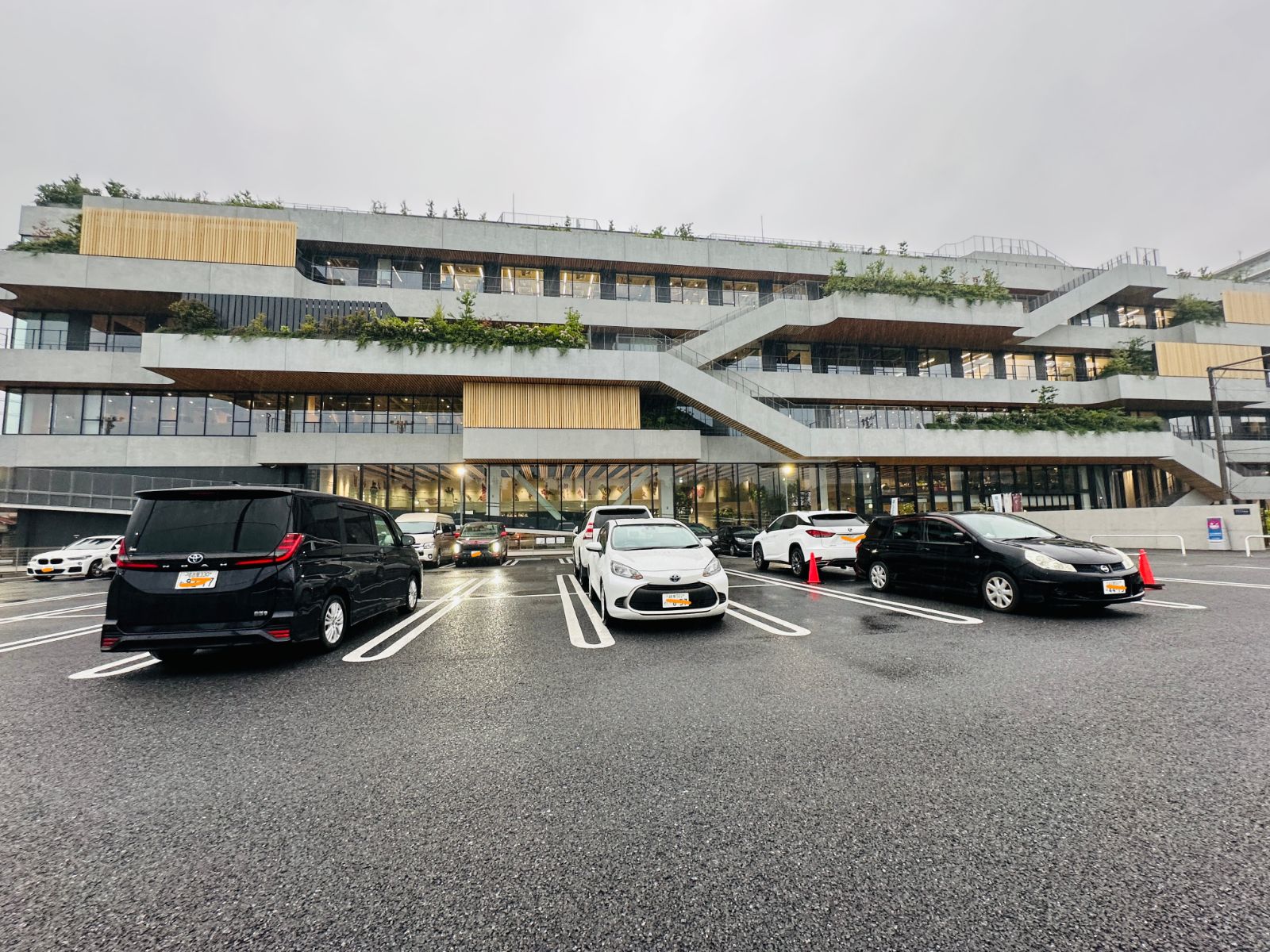陰雨天的旅館外觀，三層樓的清水模建築有階梯狀的露臺與綠色植栽，前方停車場停著幾輛車