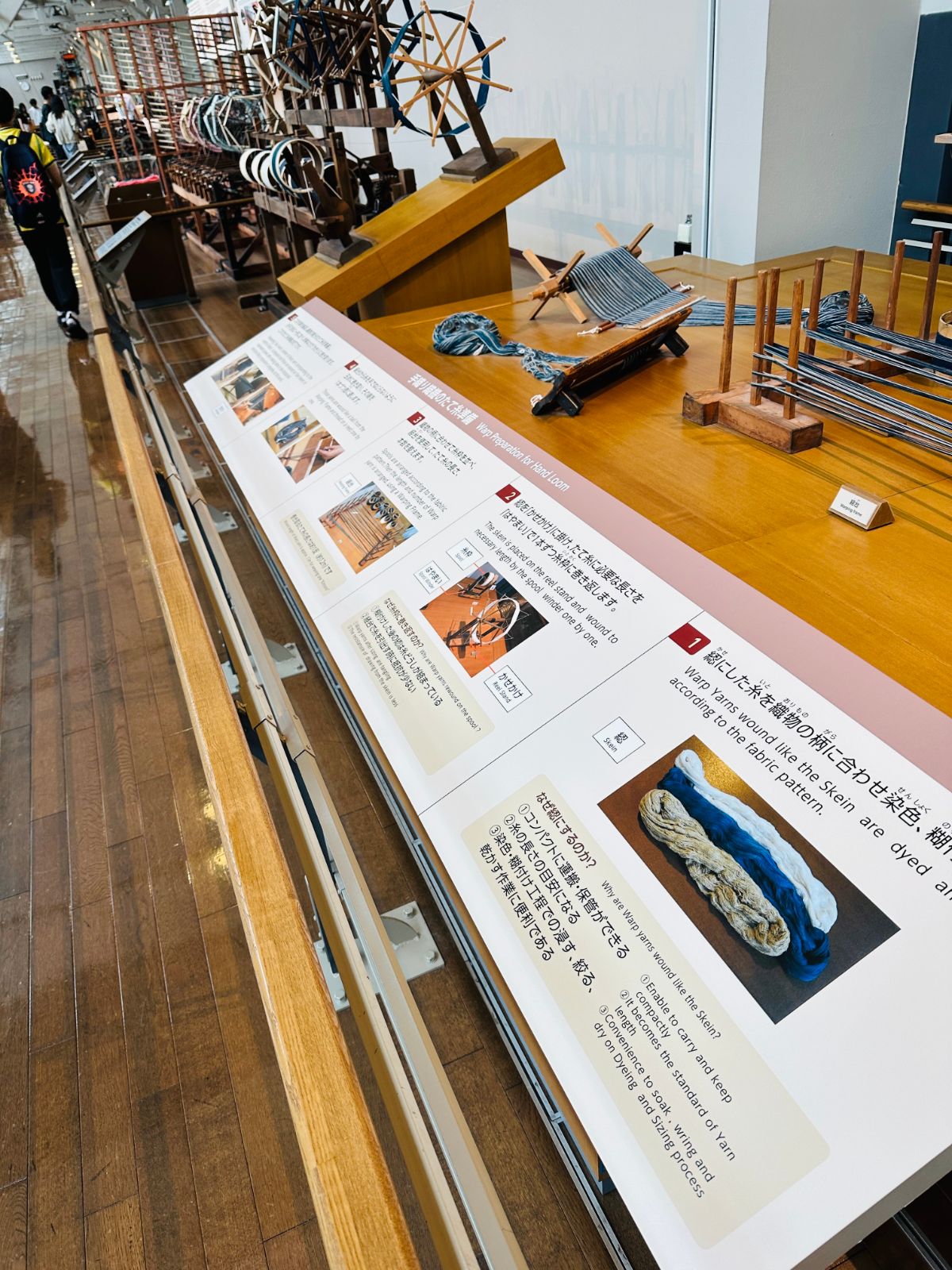 紡織工序的解說展示台，長桌上排著圖文說明板與染成藍白色的紗束，後方是木造的紡車