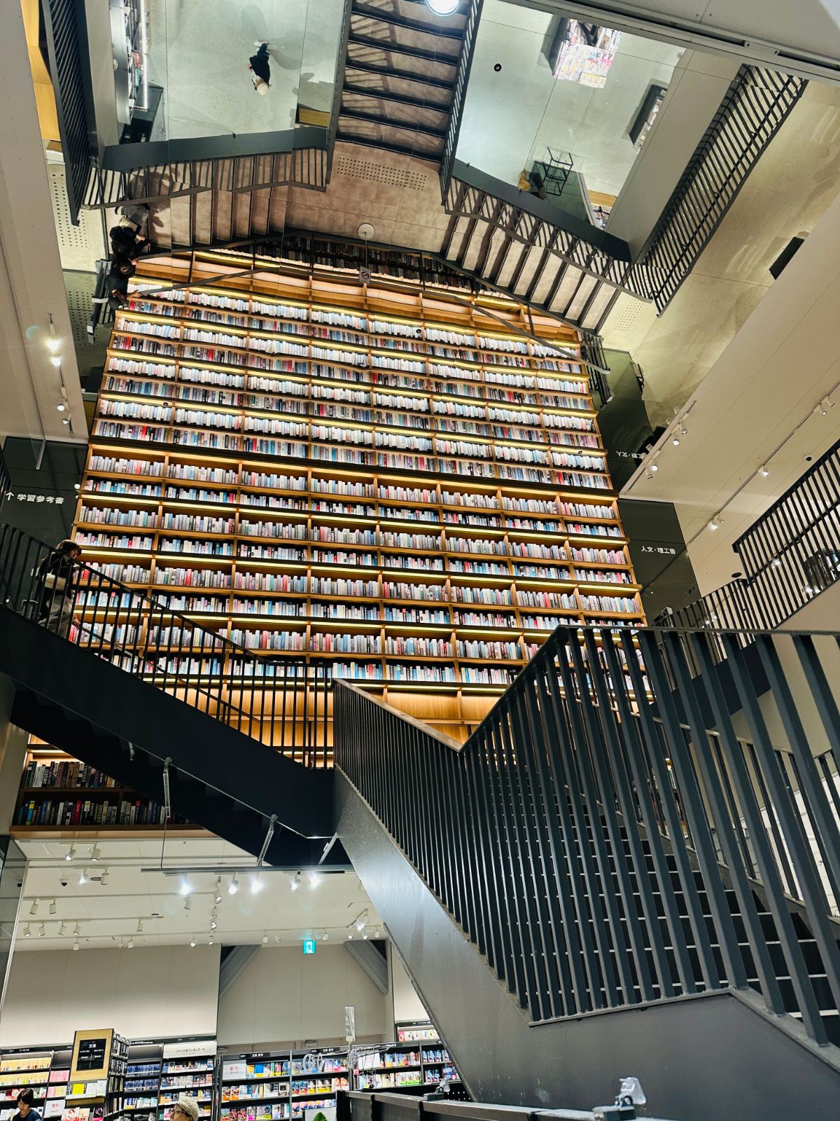 書店的挑高書牆，木色層架從地面直達天花板排滿書籍，前方是黑色鋼構的樓梯與欄杆