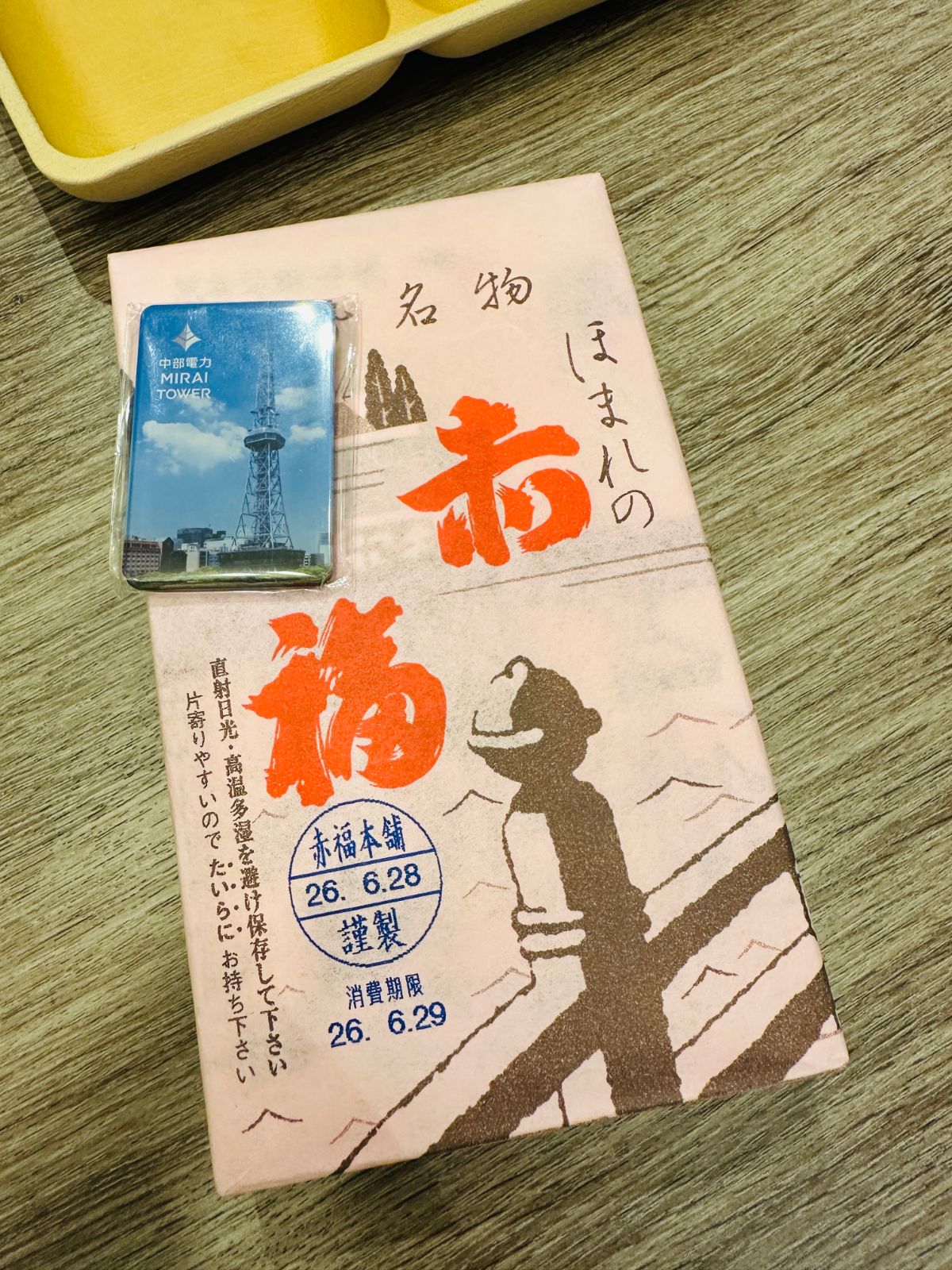 桌上放著一盒赤福，粉色包裝紙印著紅色「赤福」二字與挑扁擔的人物圖案，旁邊是 MIRAI TOWER 的磁鐵紀念品