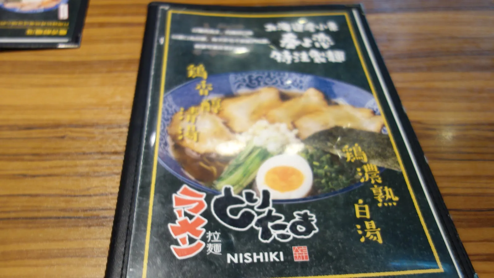 木桌上立著雞玉錦拉麵的菜單封面，深色底印著一碗拉麵的照片與「雞濃熟白湯」字樣