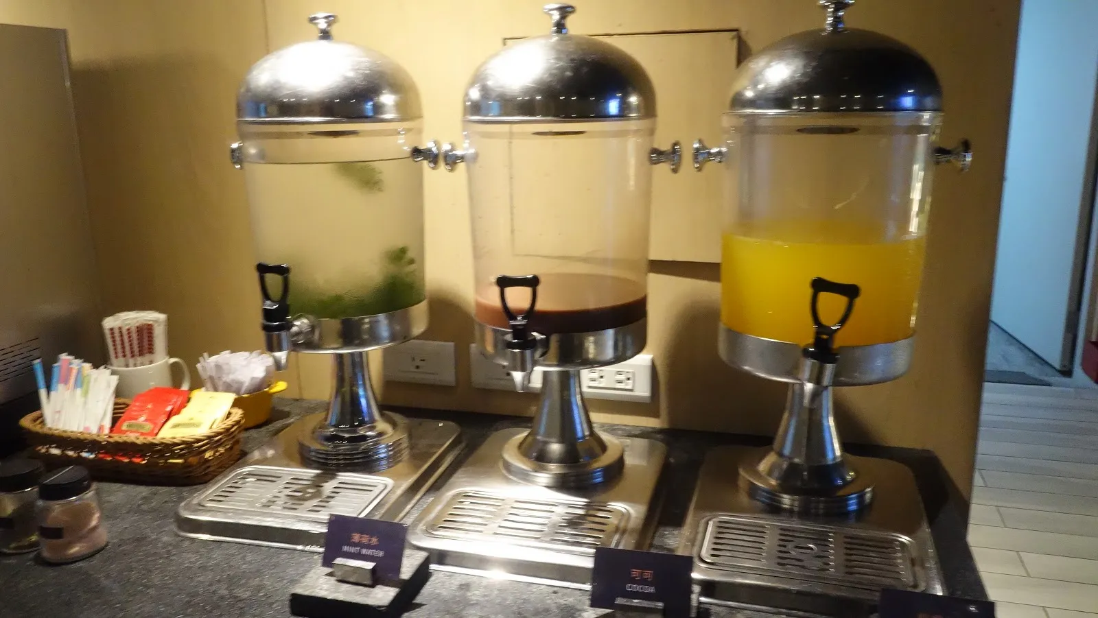 自助飲料吧，三個透明果汁桶分別裝著水、檸檬水與柳橙汁，前方籃子裡放著茶包與攪拌棒
