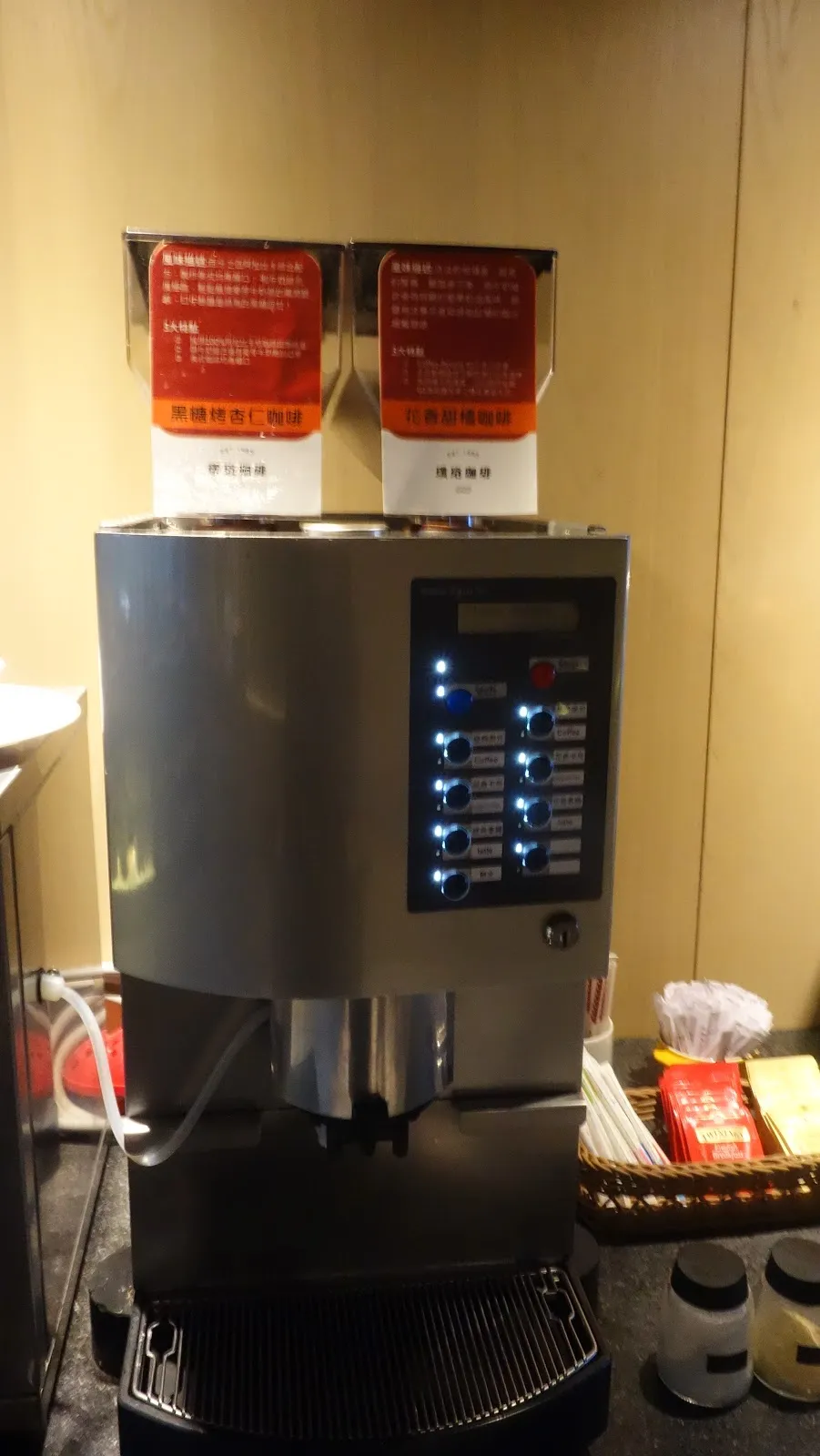 一台大型自動飲料機，機身貼著兩張紅色的操作標示，旁邊放著紙杯與濾掛包