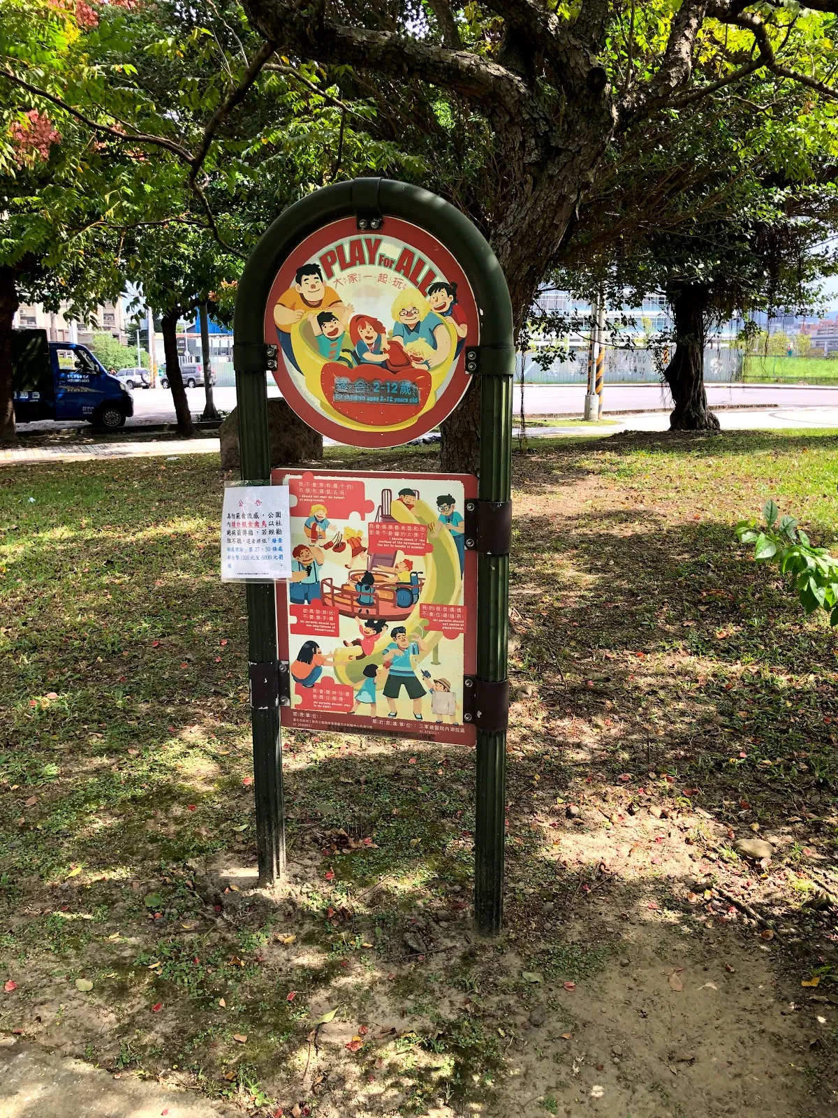 公園裡的遊戲區告示牌，圓形招牌上寫著 PLAY，下方看板印著遊戲規則插圖，後方是大樹與草地