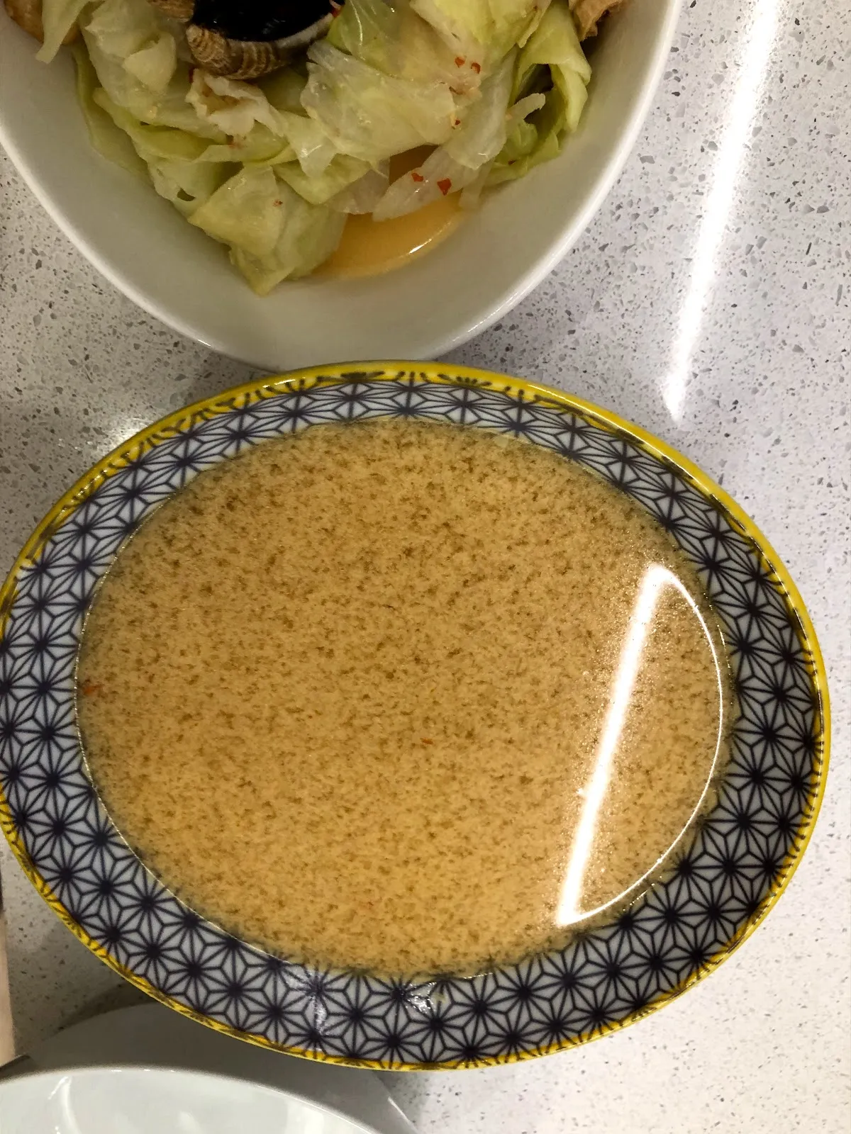 幾何花紋的碗裡盛著淺褐色的湯，後方是一碗高麗菜與蛤蜊