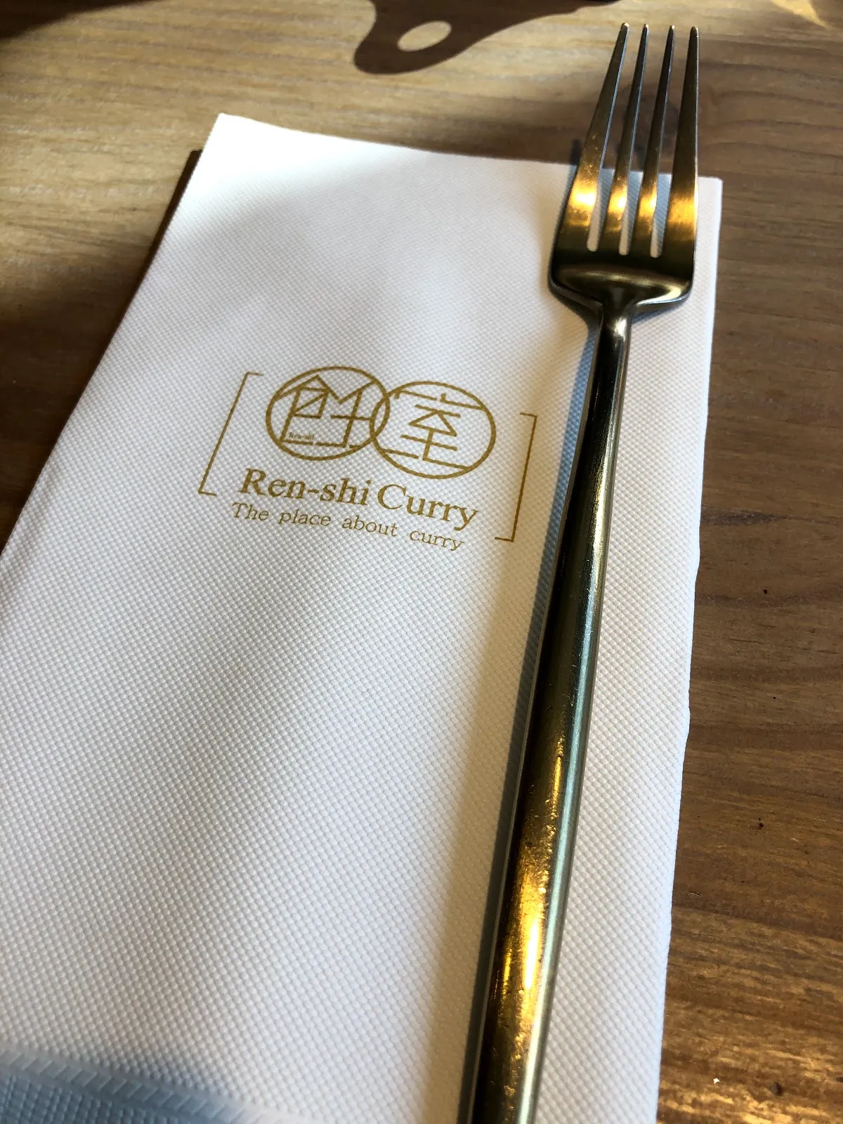 白色餐巾上印著飪室咖哩 Ren-shi Curry 的金色標誌，旁邊放著一支叉子