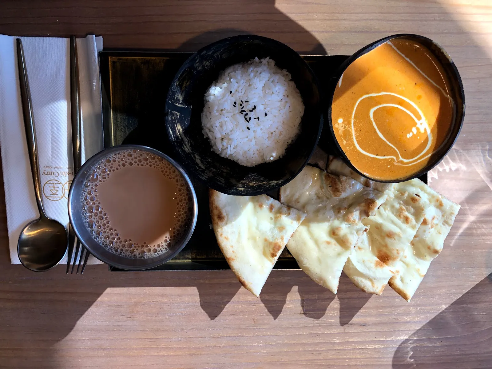 木桌上的咖哩套餐俯拍，黑盤裡是白飯、橘紅色咖哩、切片烤餅與一杯印度奶茶，旁邊放著餐具