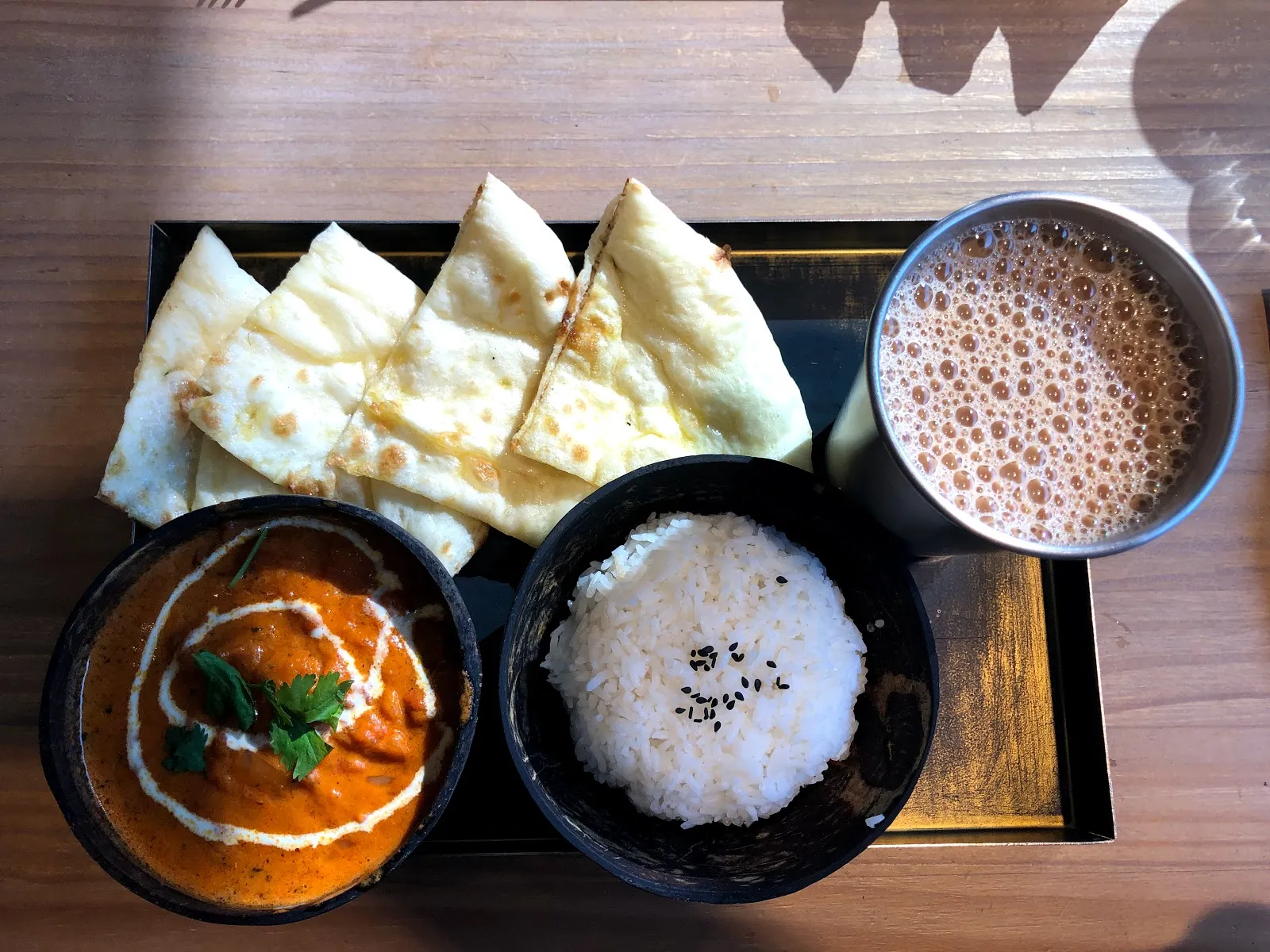 木桌上的咖哩套餐，黑色餐盤裡有一碗咖哩、一碗白飯、切片烤餅與一杯印度奶茶