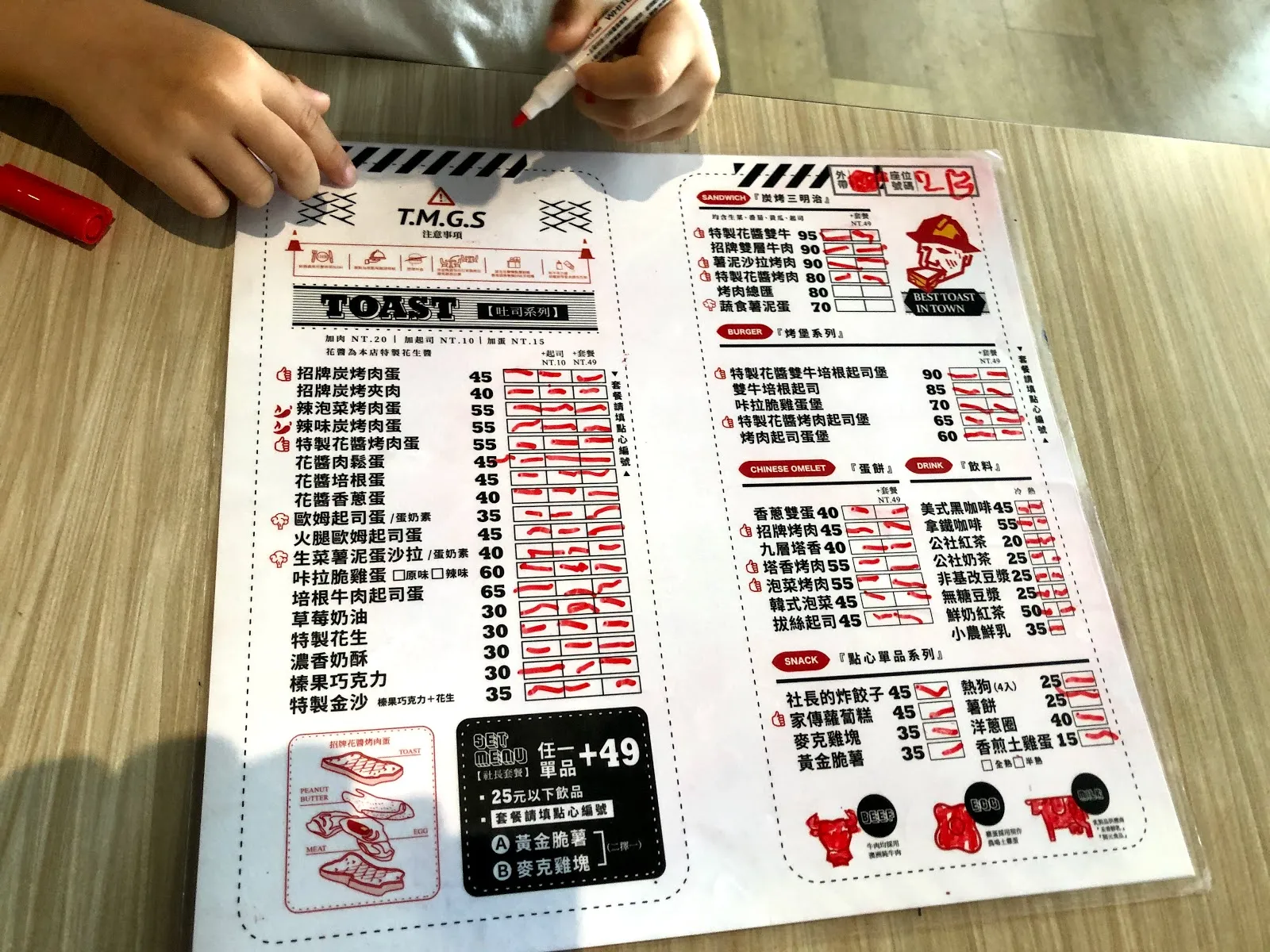 桌上攤開的 T.M.G.S 點餐單，紅框分成吐司、蛋餅與飲料等區塊，一隻手正拿著筆勾選