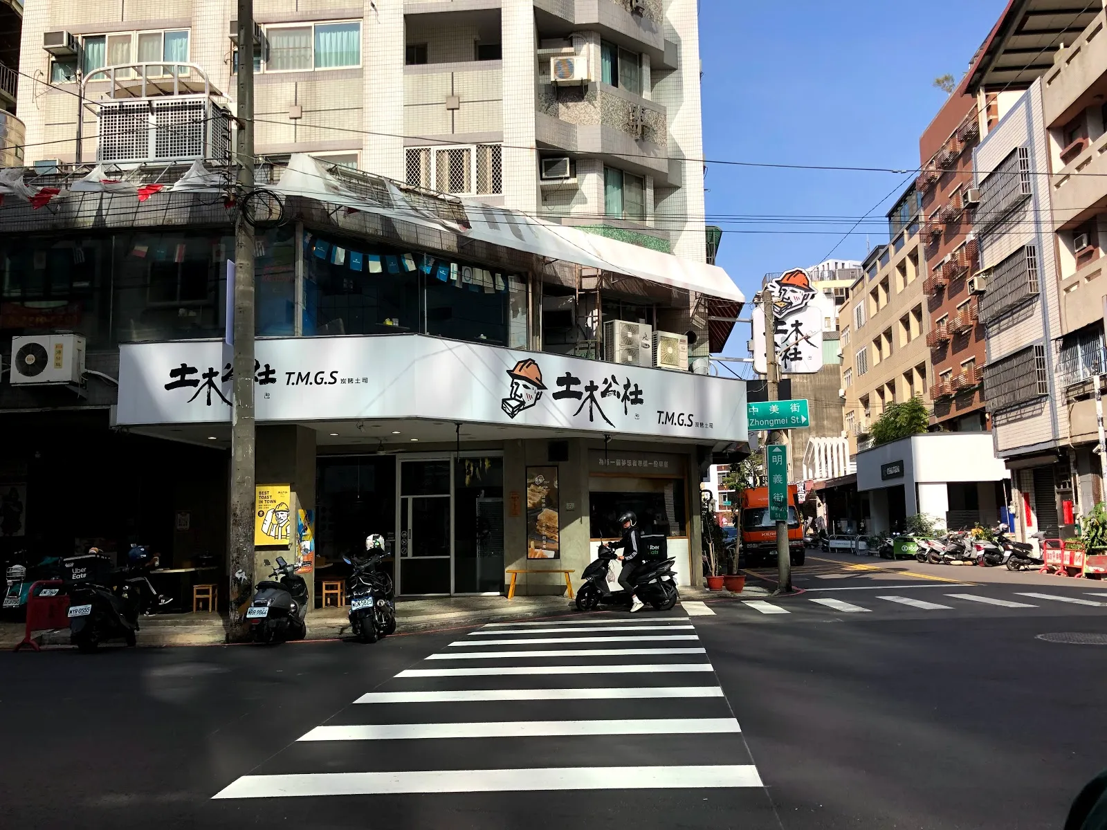 土木公社的店門外街景，街角的白色店面掛著 T.M.G.S 招牌，前方是斑馬線與公寓大樓