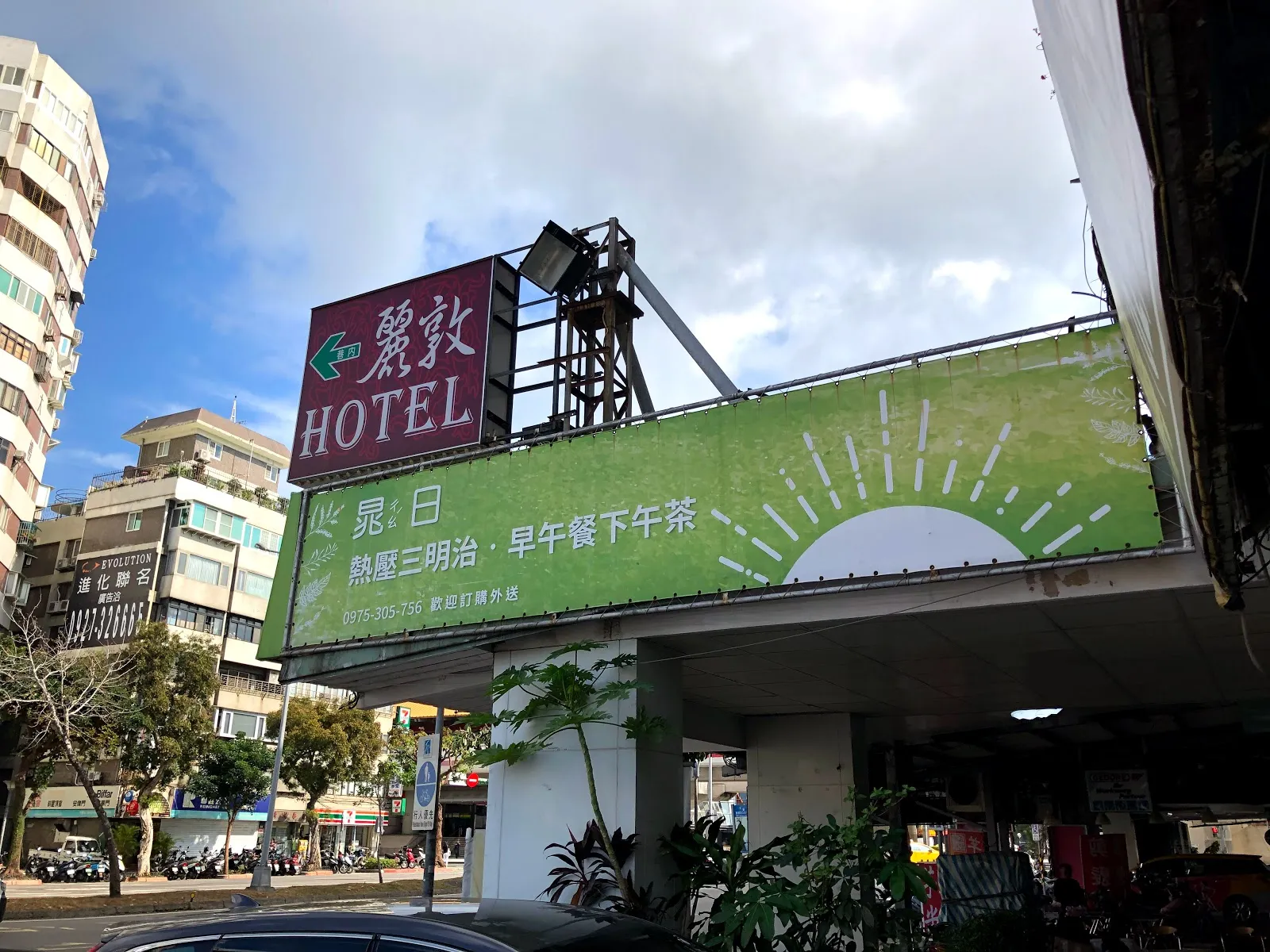 街邊仰角，綠色的廣告看板寫著熱壓三明治與早午餐、下午茶，後方是旅館招牌與公寓大樓