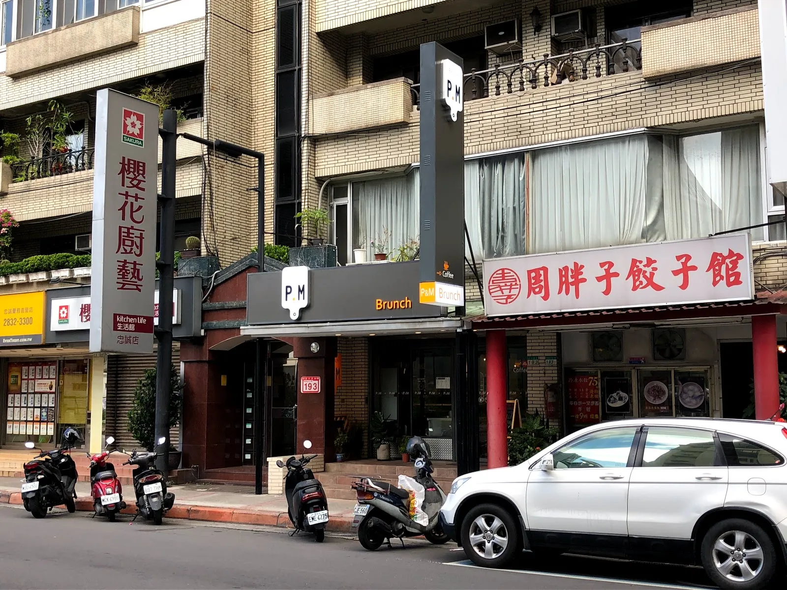 店家所在的街景，騎樓下並排著 P.M Brunch、櫻花廚藝與周胖子餃子館的招牌，路邊停著機車與白色休旅車