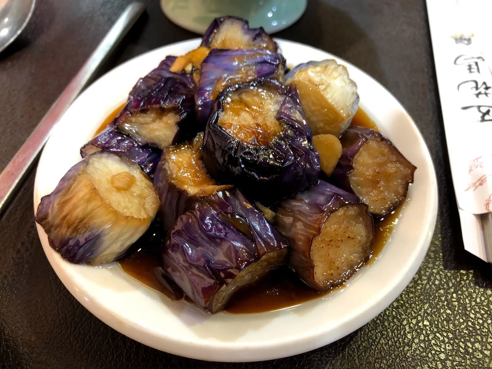 白色圓盤上的紅燒茄子，茄段淋著深褐色醬汁與蒜末