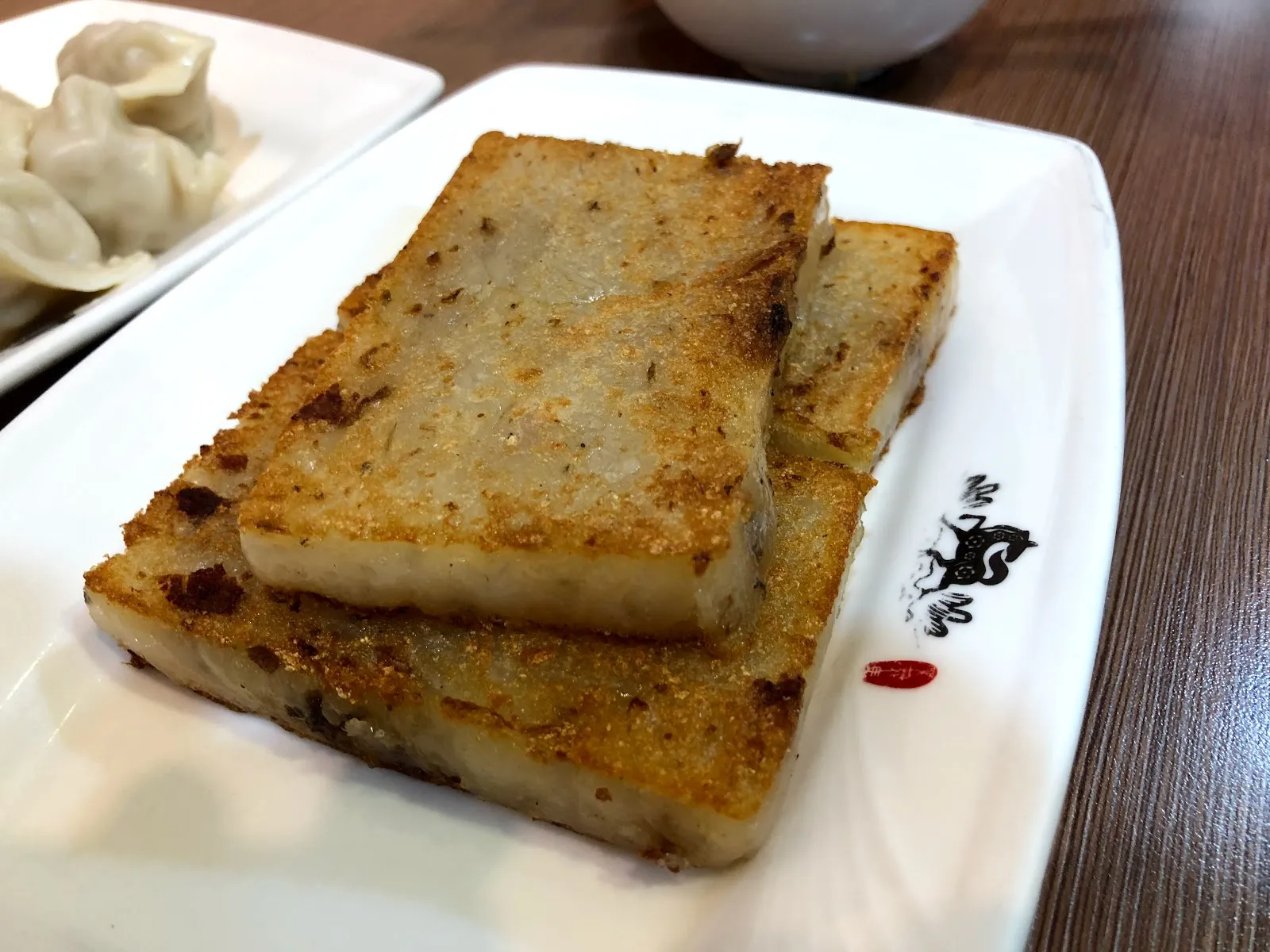 白色長盤上的兩塊煎蘿蔔糕，表面煎得金黃，後方是一盤蒸餃