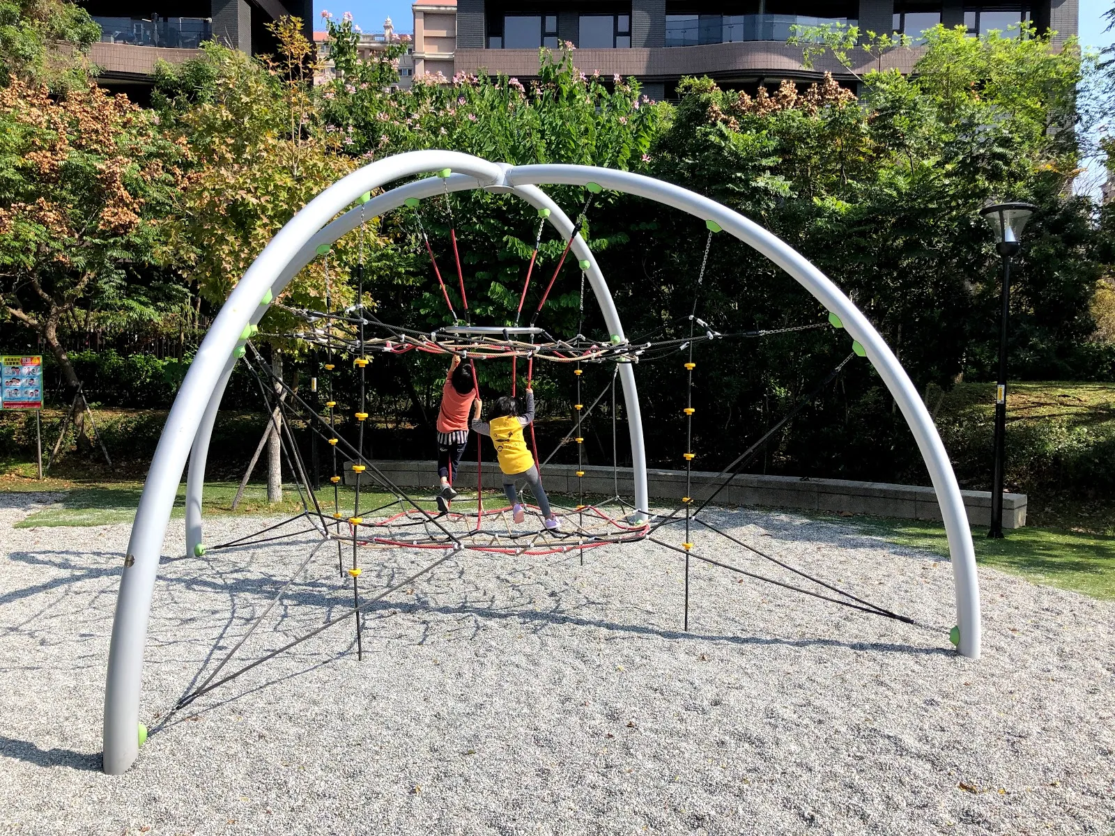 公園裡的大型攀爬架，白色拱形鋼架吊著紅色繩網，一個小孩正爬在網子中央，背景是綠樹與住宅