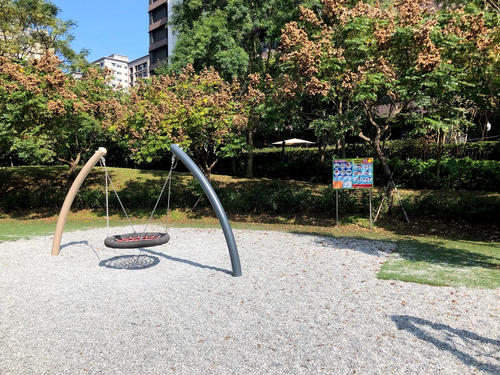 公園的鳥巢式盪鞦韆，兩根白色拱柱吊著圓形的網狀座椅，四周是碎石地與綠樹