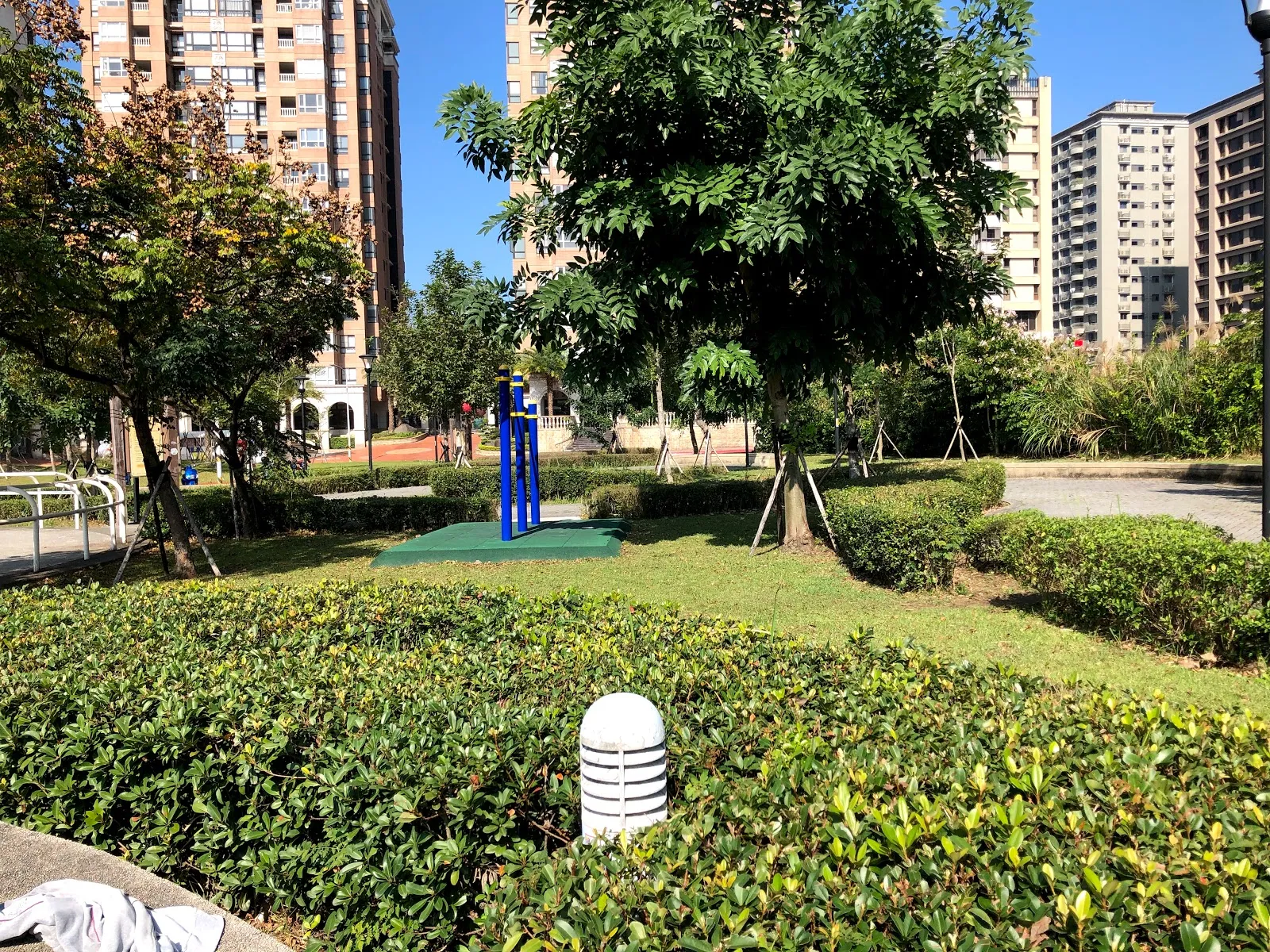 公園步道旁的綠籬與大樹，遠方是藍天下的公寓大樓，草地上放著運動器材
