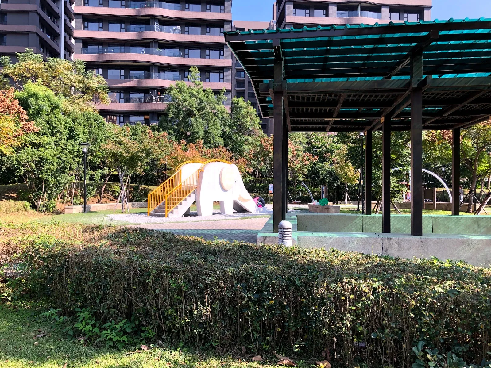 公園的遮陽棚架與大象造型的磨石子溜滑梯，前方是修齊的綠籬，後方是粉色外牆的大樓