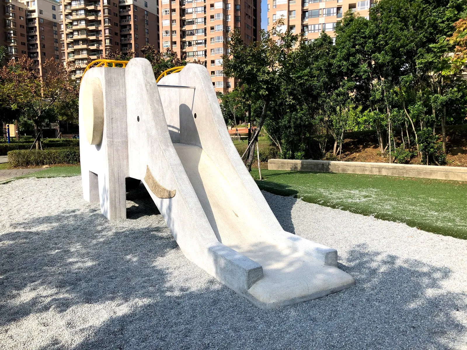大象造型的磨石子溜滑梯，象鼻形成滑道，四周鋪著碎石，旁邊是綠色人工草皮