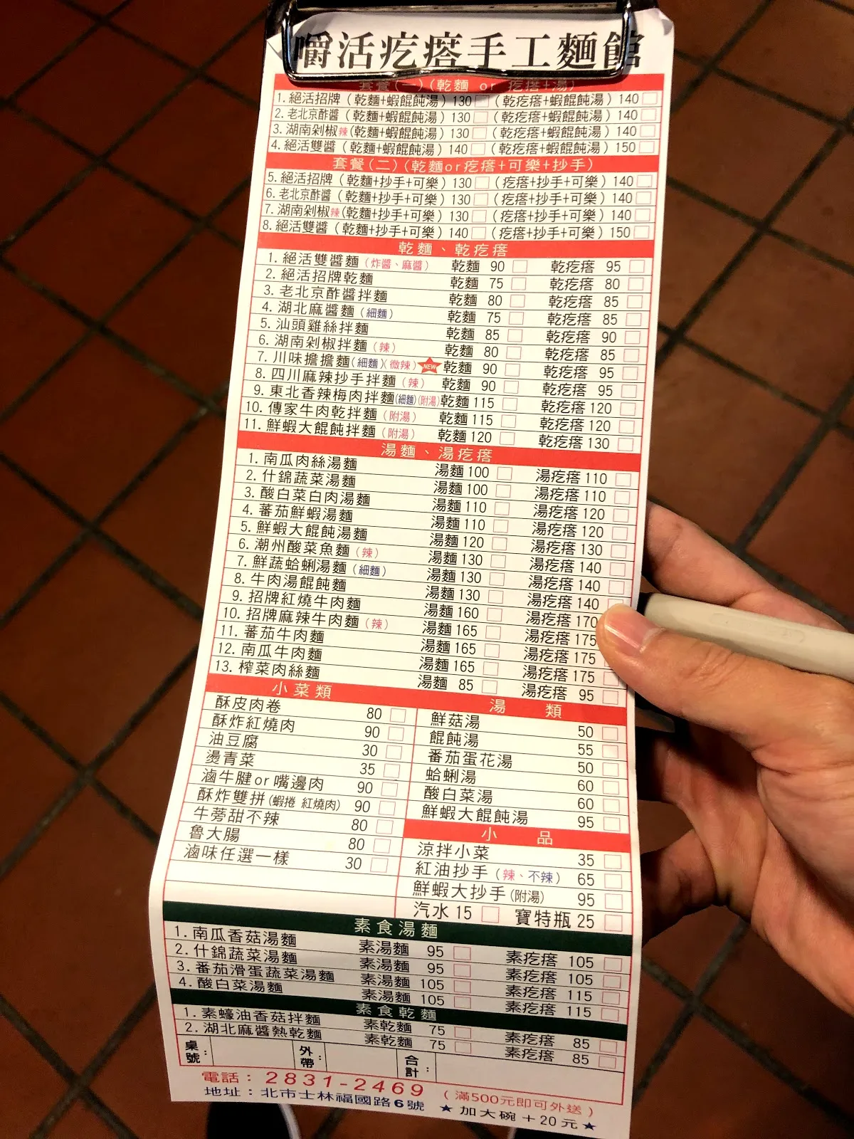 手上拿著嚼活疙瘩手工麵館的點菜單，紅框表格列滿麵食、湯品與小菜的價格，下方是士林區的地址與電話