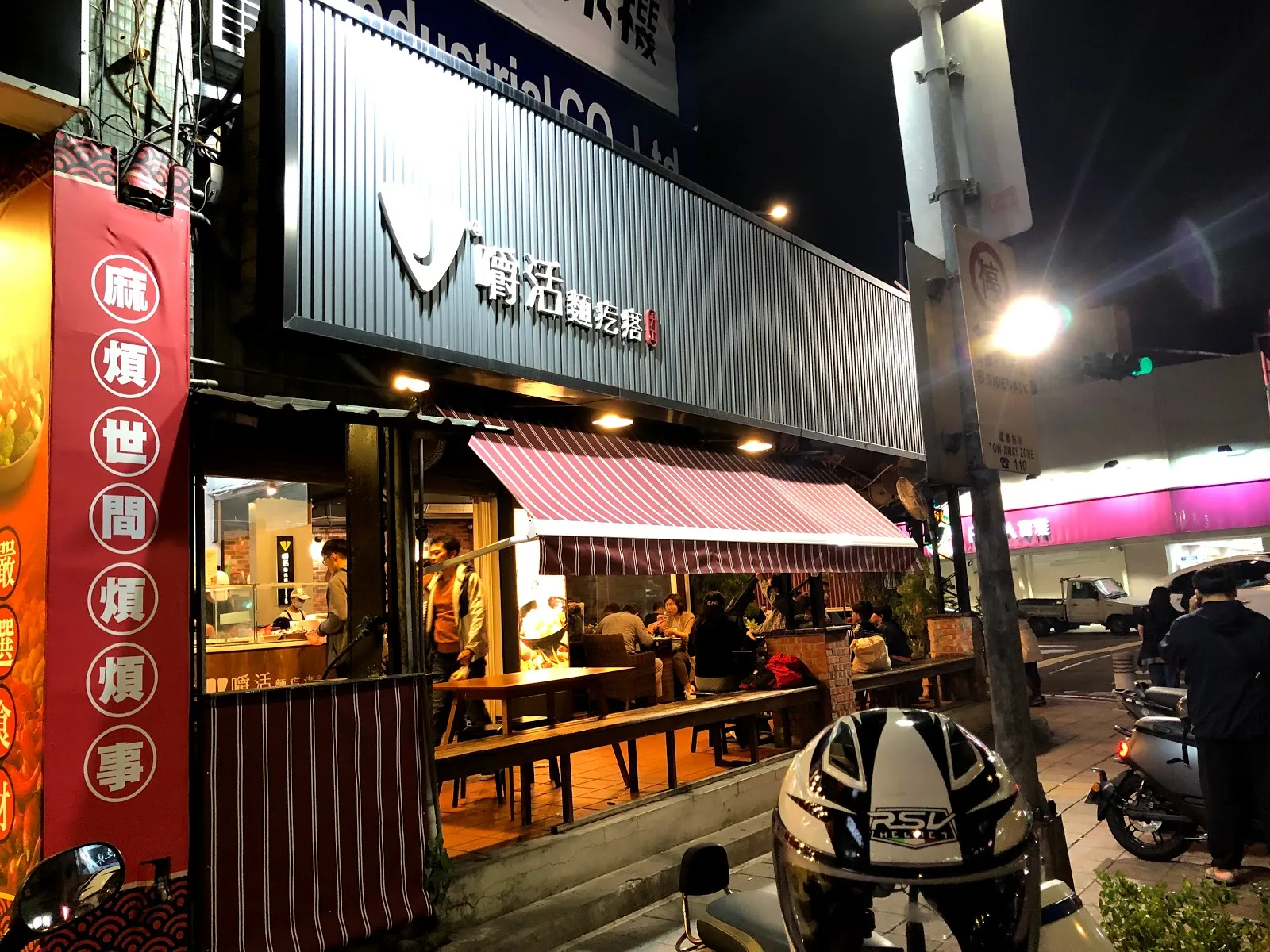 夜晚的店面，白色浪板外牆掛著招牌，門口立著紅色的直立布條，騎樓下停著機車