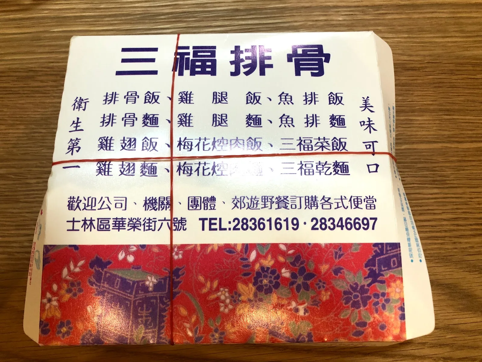三福排骨的紙製便當盒蓋，藍字印著排骨飯、雞腿飯、魚排飯等品項，下方是士林區華榮街的地址與電話