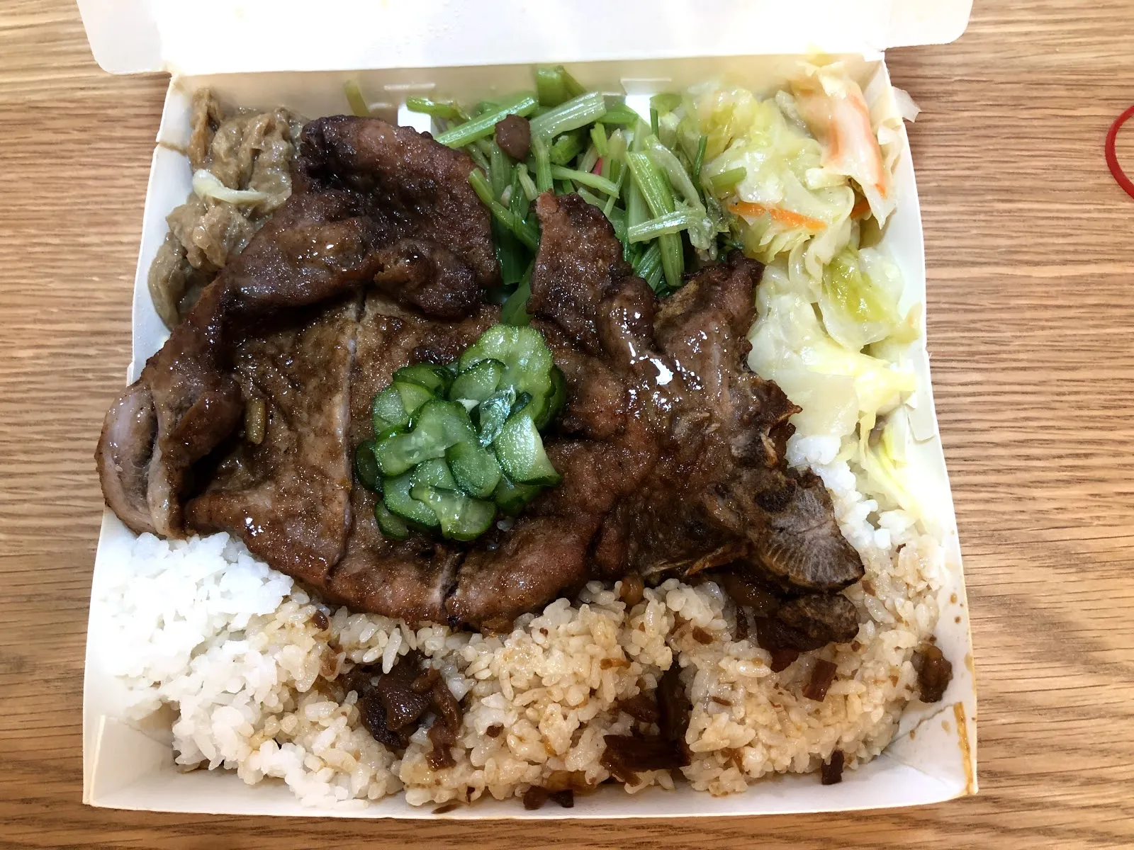 紙盒便當，白飯上鋪著醬色的排骨肉片與醃黃瓜，旁邊配著炒青菜與高麗菜