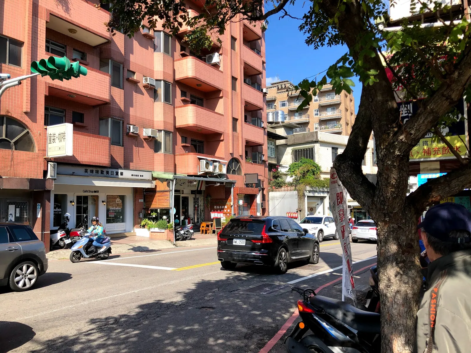 白天的街景，行道樹下停著汽車與機車，對面是紅磚色的公寓樓