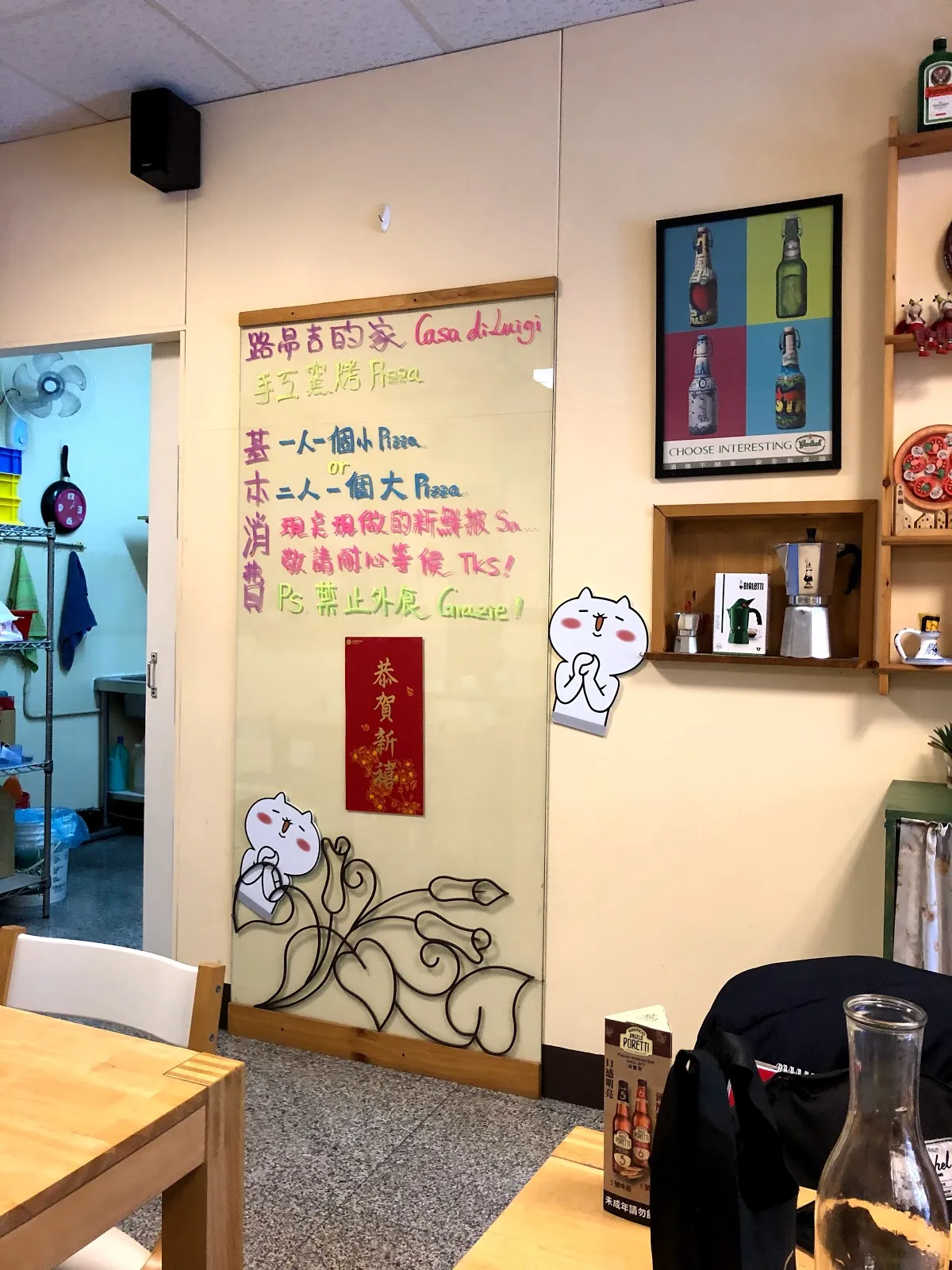 店內一角，牆上用彩色手寫字寫著店名與座位說明，木桌旁擺著長凳，牆架上掛著平底鍋