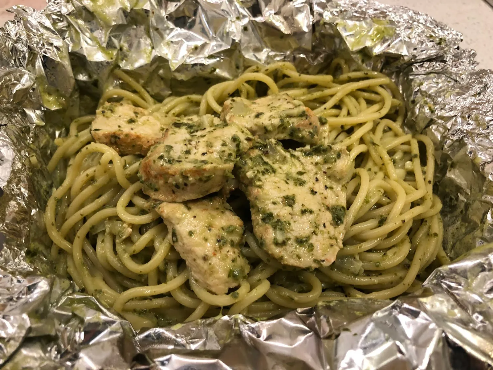 鋁箔盒裡的青醬義大利麵,麵條裹著綠色青醬,上面鋪著切片的雞胸肉與香草碎
