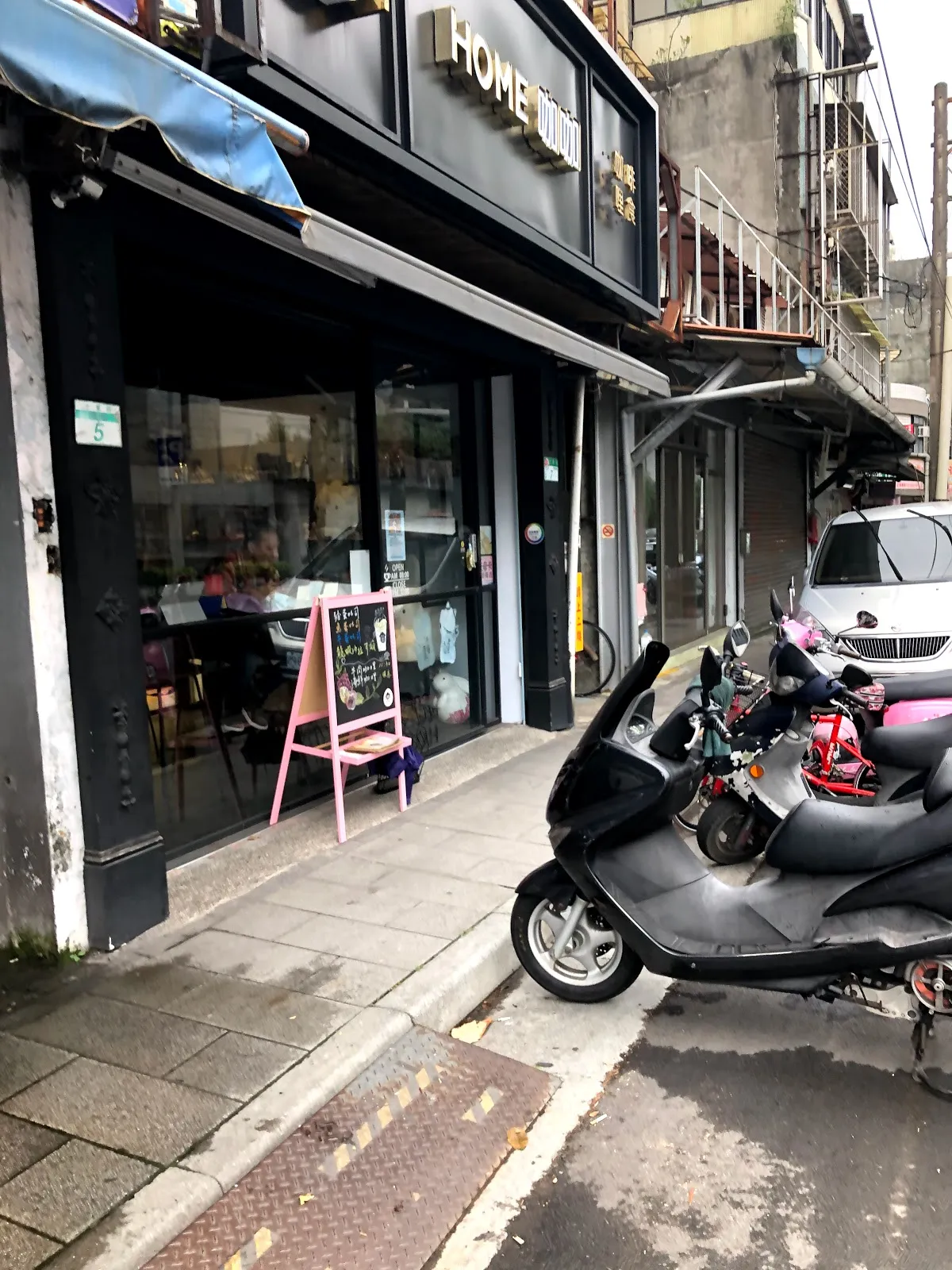 HOME 咖咖的店門外巷弄街景，黑色門面掛著 HOME 招牌，門口立著粉紅色的立式菜單牌，路邊停著機車