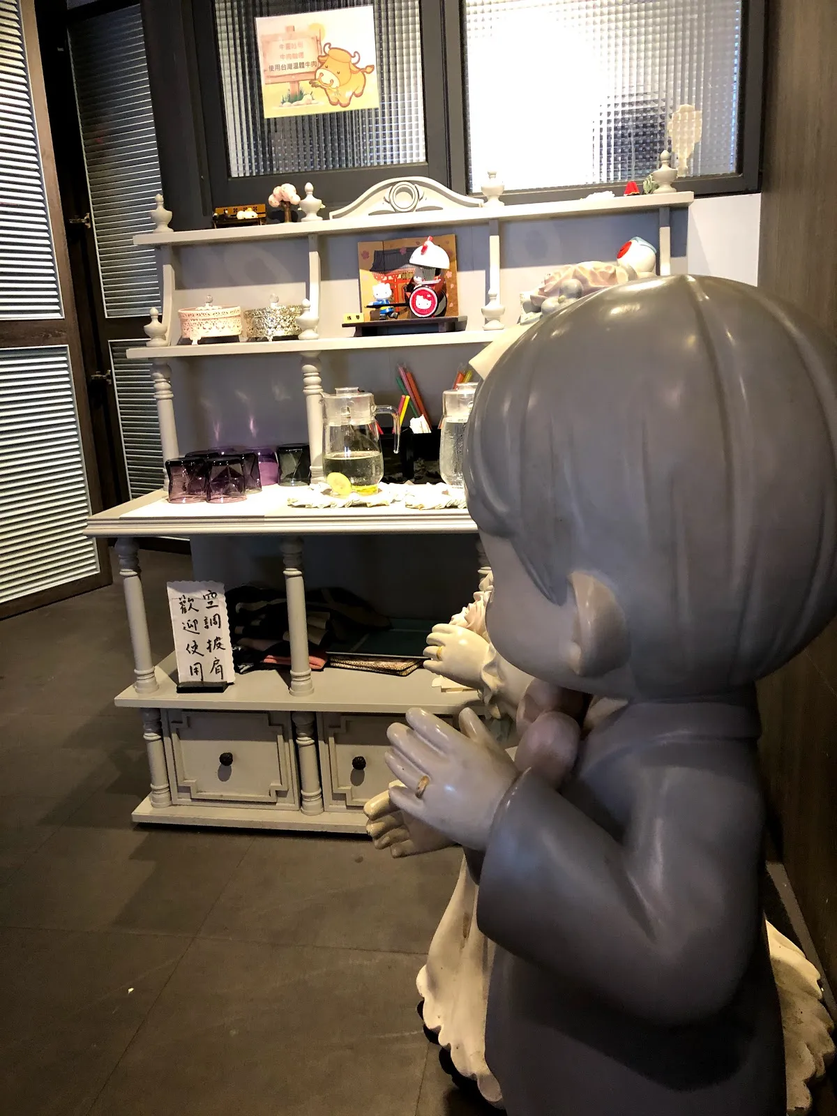 店內一角，一尊灰色的女孩雕像雙手合在胸前，後方的白色層架上擺著相框與玻璃罐