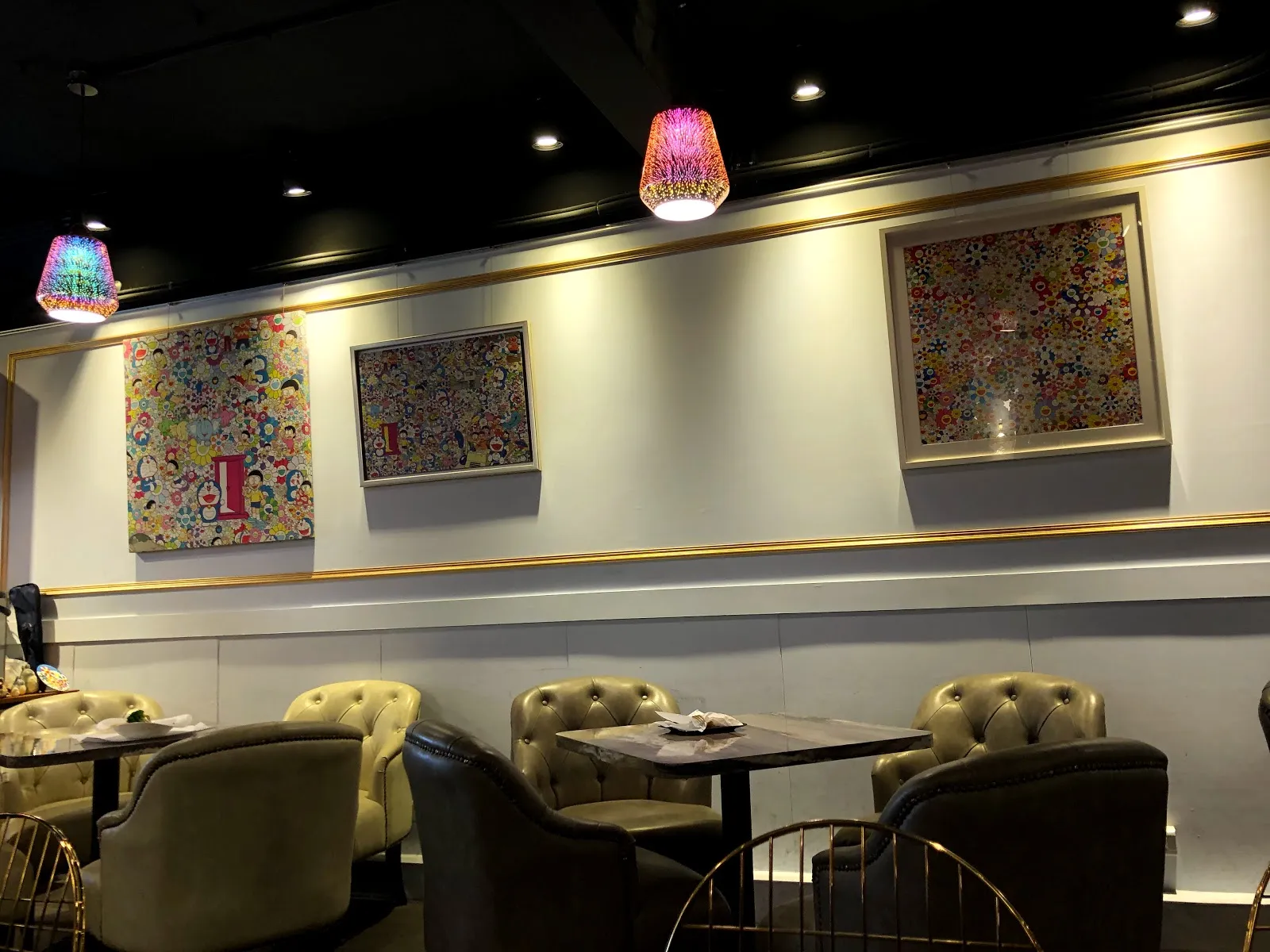 店內用餐區，米色絨布沙發椅配深色圓桌，牆上掛著三幅色彩繽紛的畫作，天花板垂著花紋燈罩