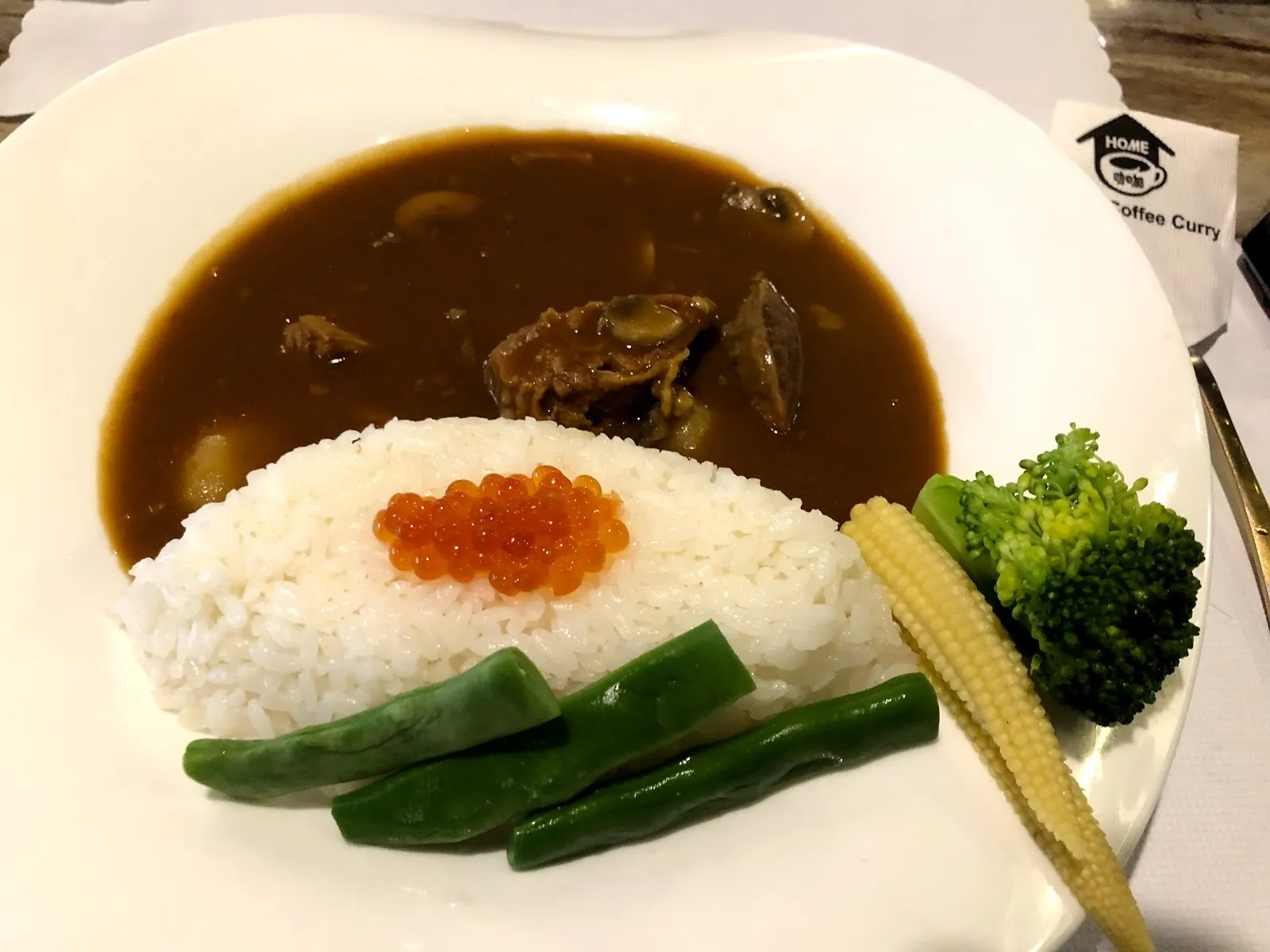 白色盤子上的咖哩飯，白飯中央點著橘紅色的鮭魚卵，深褐色咖哩醬淋在一旁，配上綠花椰菜、玉米筍與碗豆莢