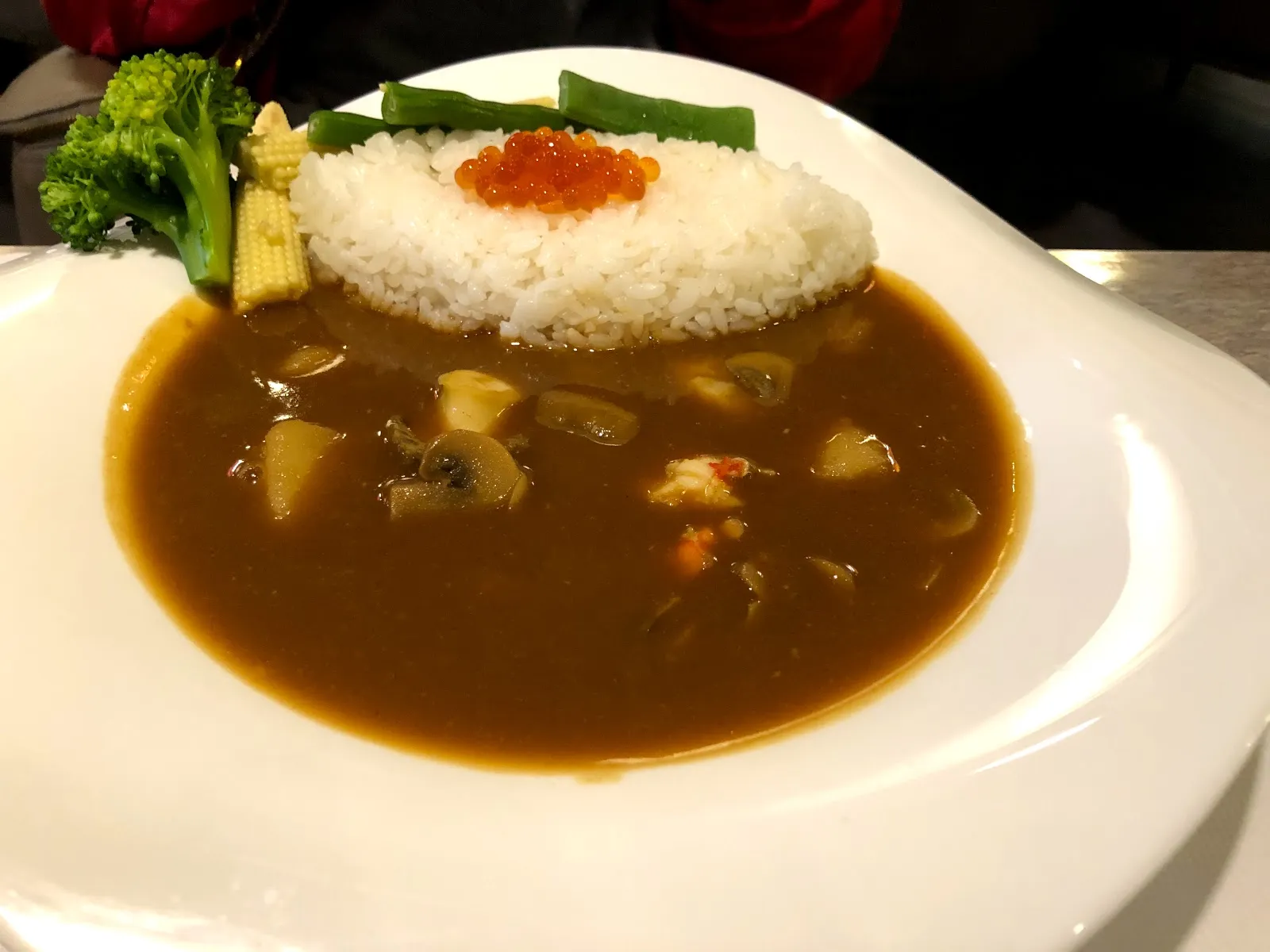 白盤上的海鮮咖哩飯，咖哩醬裡有蛤蜊與蝦仁，白飯上點著鮭魚卵，旁邊配綠花椰菜與玉米筍