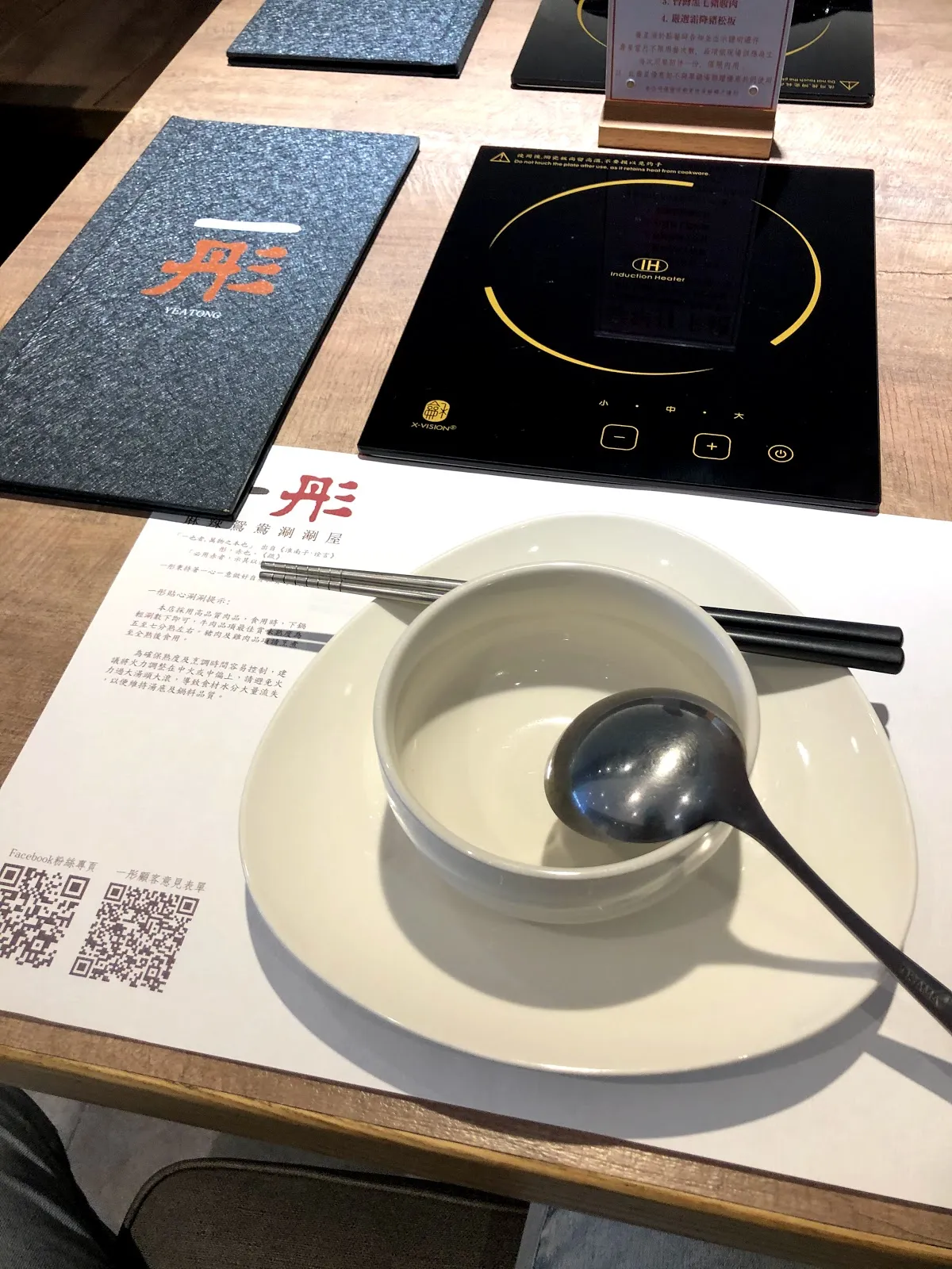 一人一鍋的座位,木桌上嵌著黑色電磁爐,旁邊放著菜單、白色沾醬碟與湯匙