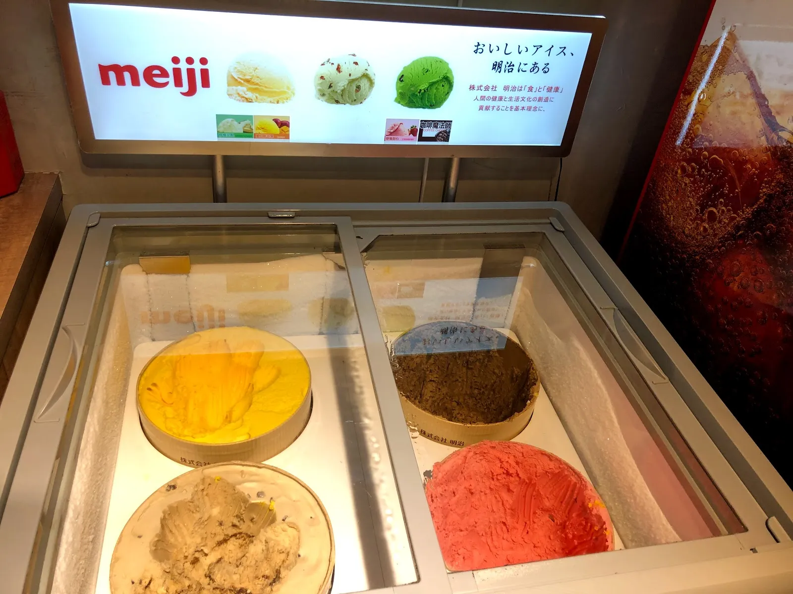 冰淇淋冷凍櫃,玻璃蓋下擺著黃色、褐色與粉紅色三桶冰淇淋,後方立著 meiji 的燈箱看板