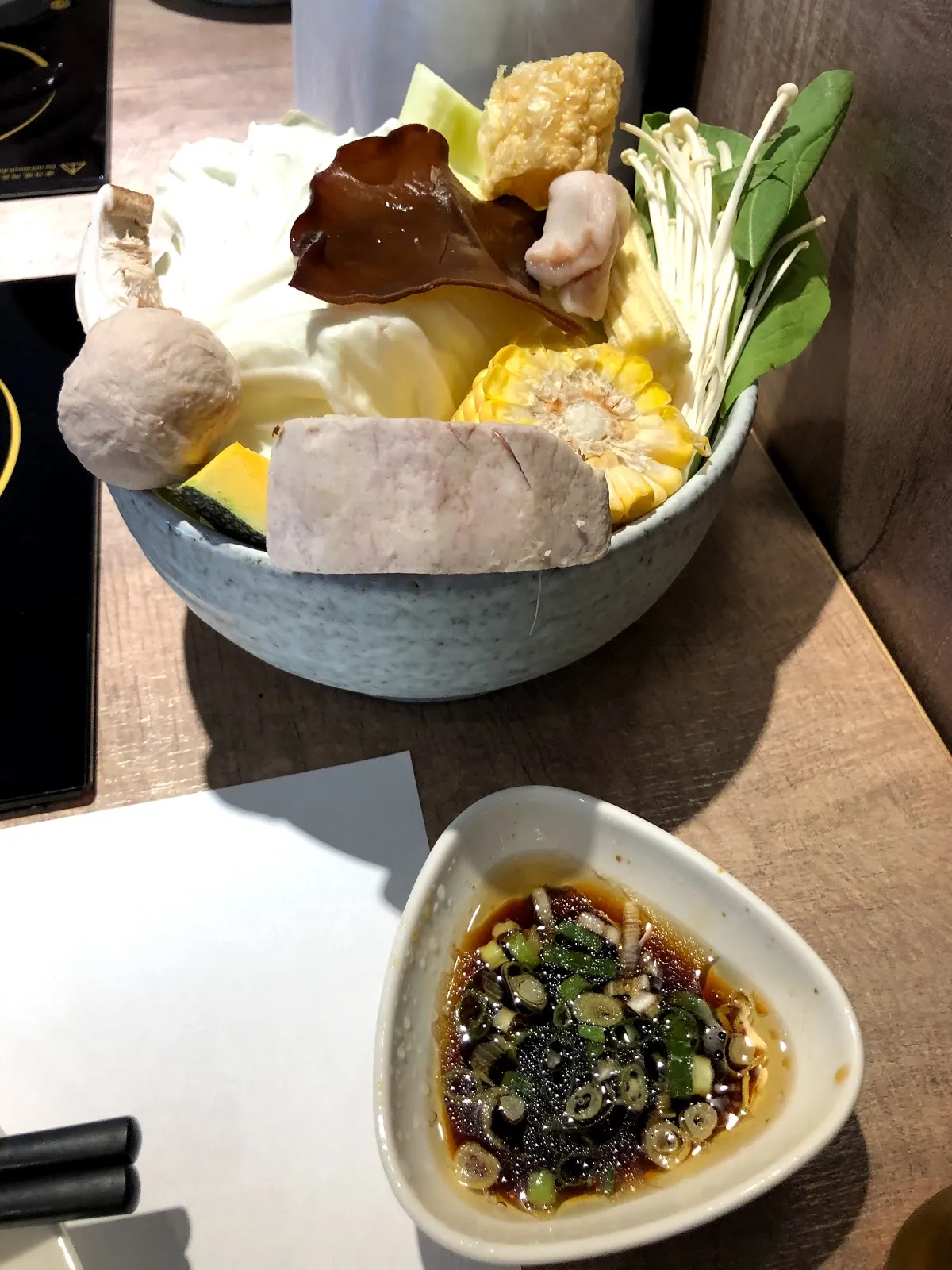 灰色陶碗盛的火鍋菜盤,有玉米、木耳、豆皮、芋頭、菇類與蔬菜,前方是一碟撒了蔥花的沾醬