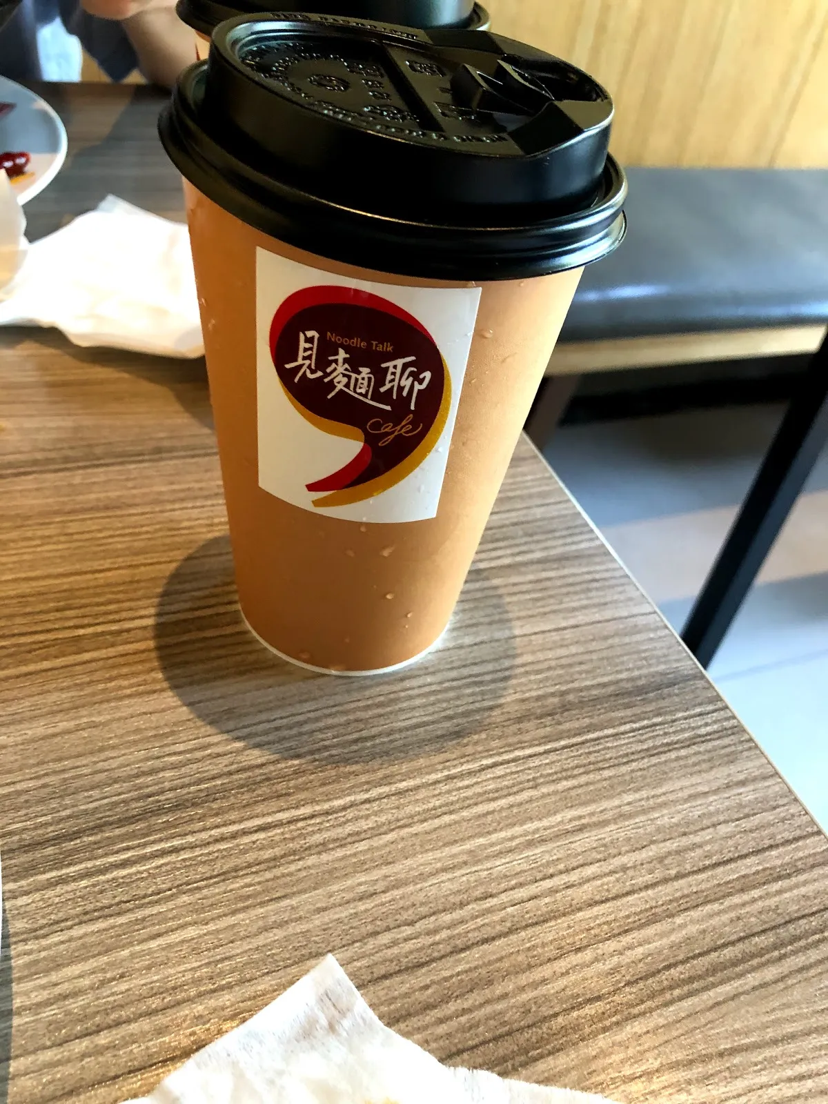木桌上的外帶飲料杯，白色紙杯貼著「見麵聊」的紅色圓形標籤，配黑色杯蓋