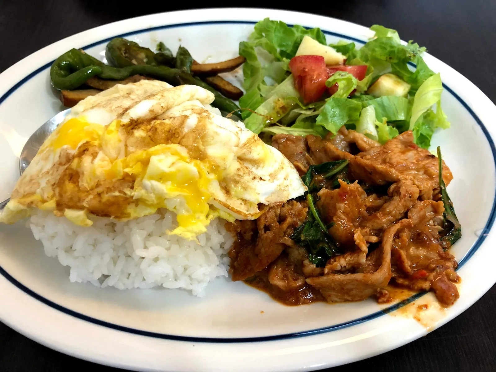 白色橢圓盤上的泰式打拋豬飯，白飯配一顆半熟荷包蛋、炒過的青椒與絞肉，旁邊是生菜番茄沙拉