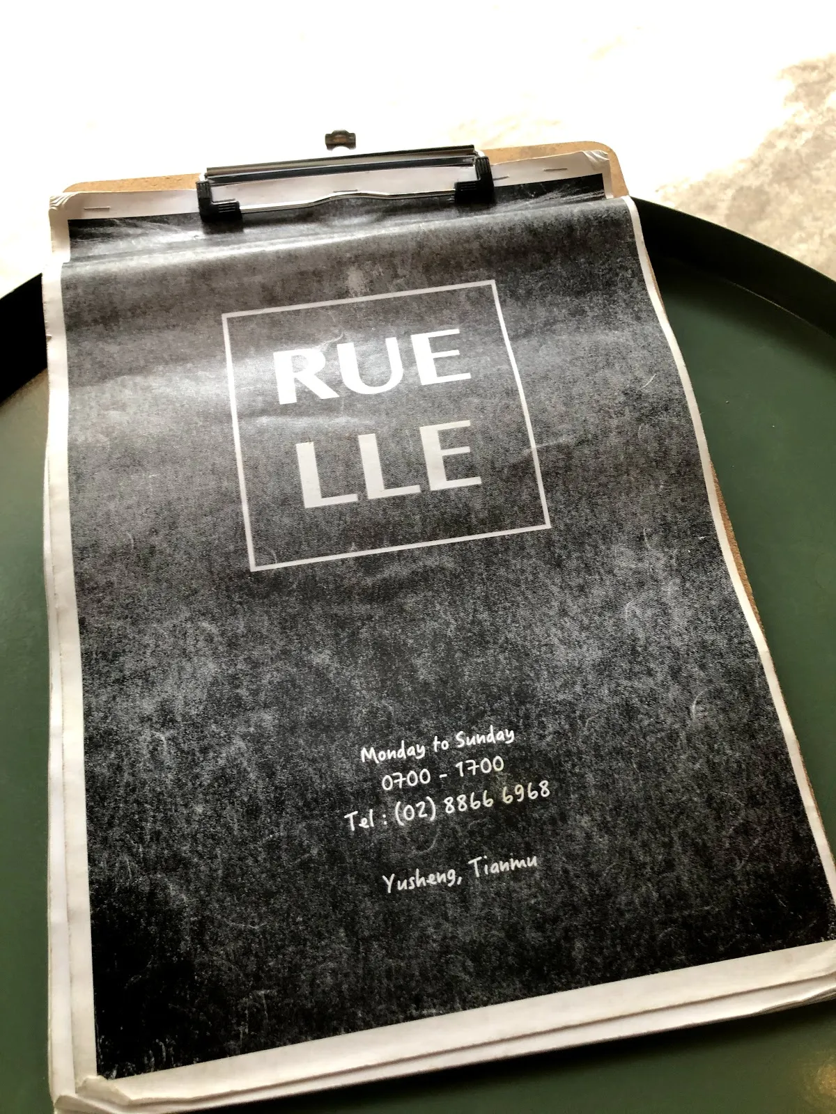 綠色圓托盤上的板夾菜單，黑色封面印著 RUELLE 的白色字樣與營業時間 09:00–17:00，地址在天母玉聖