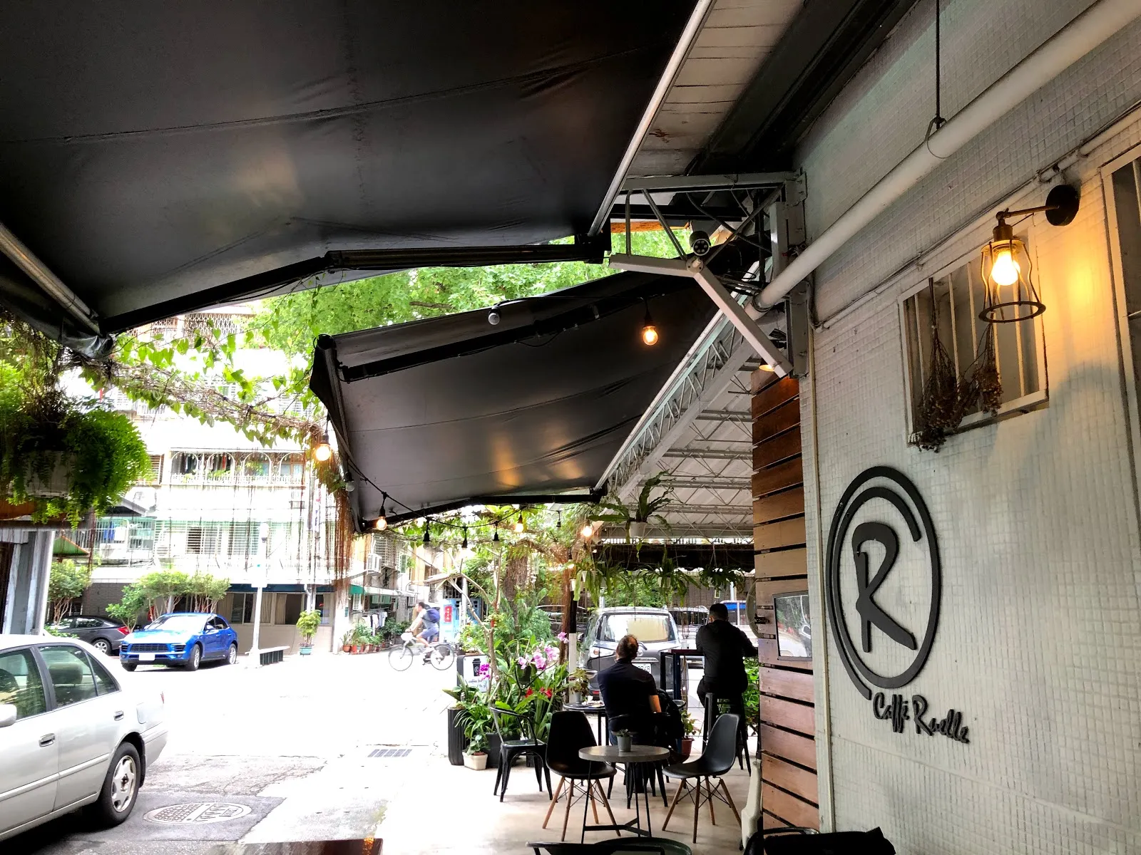 咖啡店的半戶外座位，黑色遮陽棚下擺著桌椅，牆上有大大的 R 字標誌，外面是巷弄街景與停放的車輛
