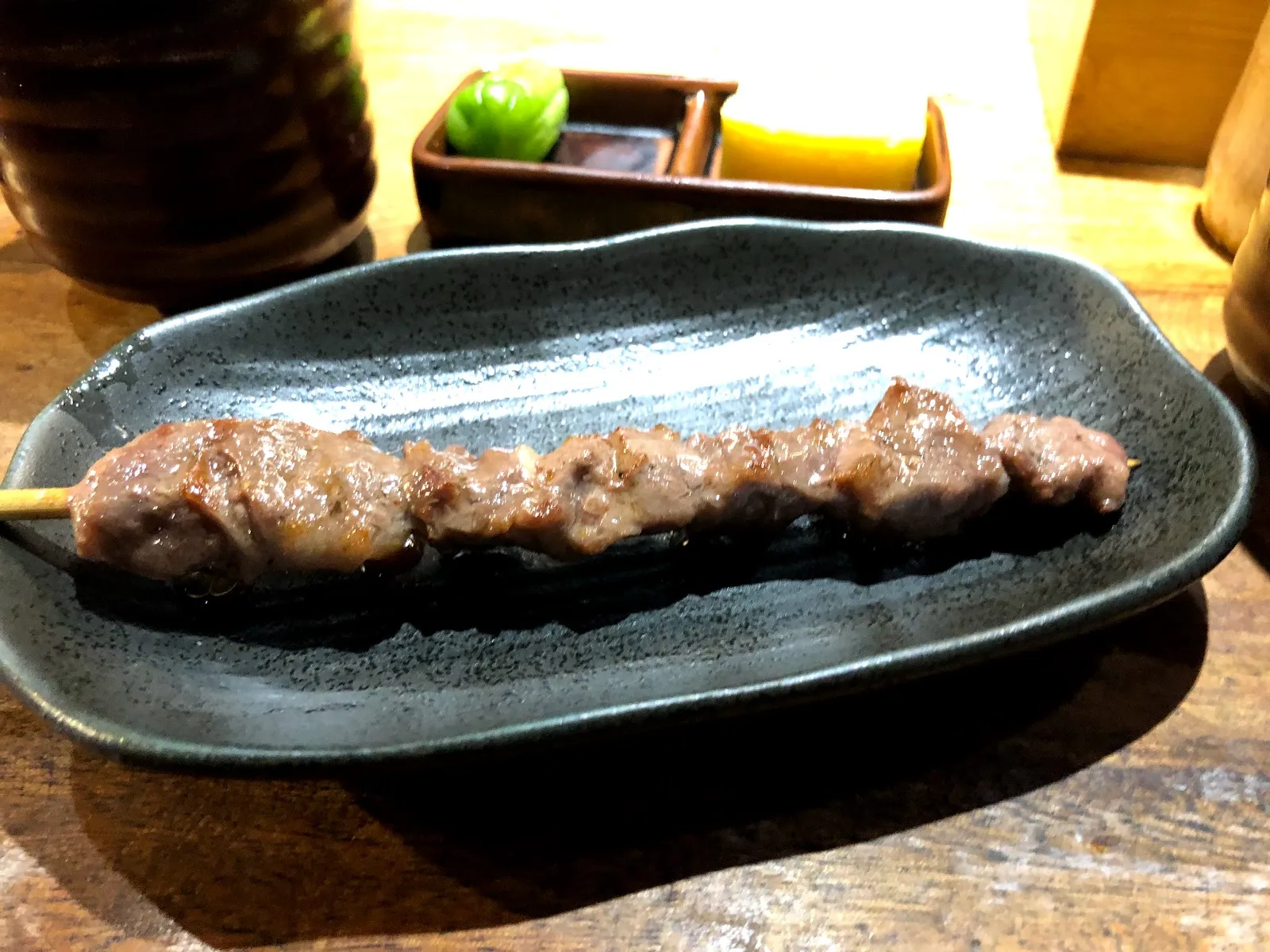 深綠色橢圓陶盤上一支烤得焦香的肉串，後方是木桌與裝著調味料的方盒