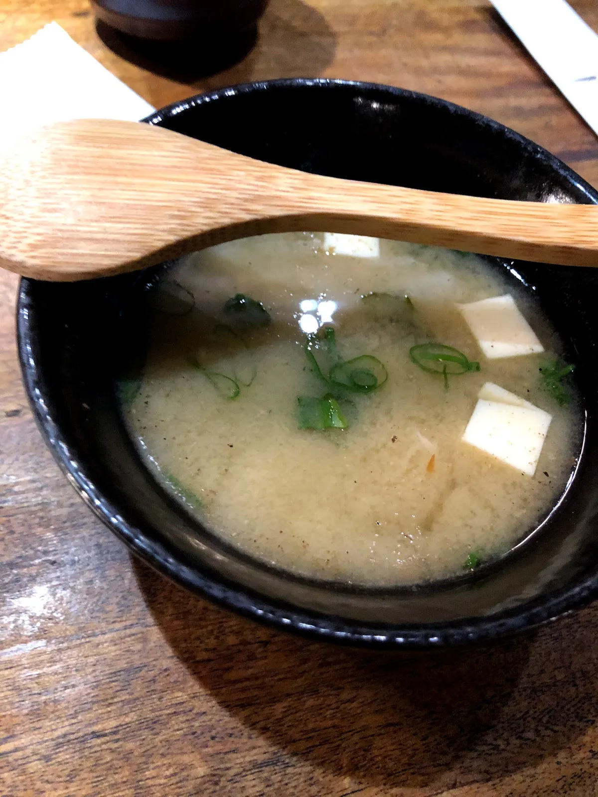 黑色小碗裡的味噌湯，湯裡浮著白色豆腐塊與蔥花，木湯匙橫放在碗口
