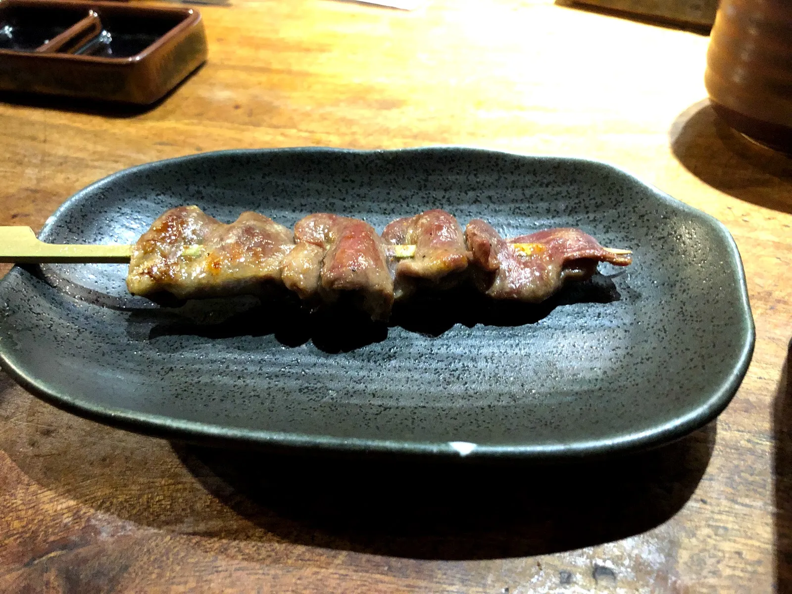 深綠色陶盤上的雞肉串燒，四塊肉之間夾著蔥段，肉色偏粉紅